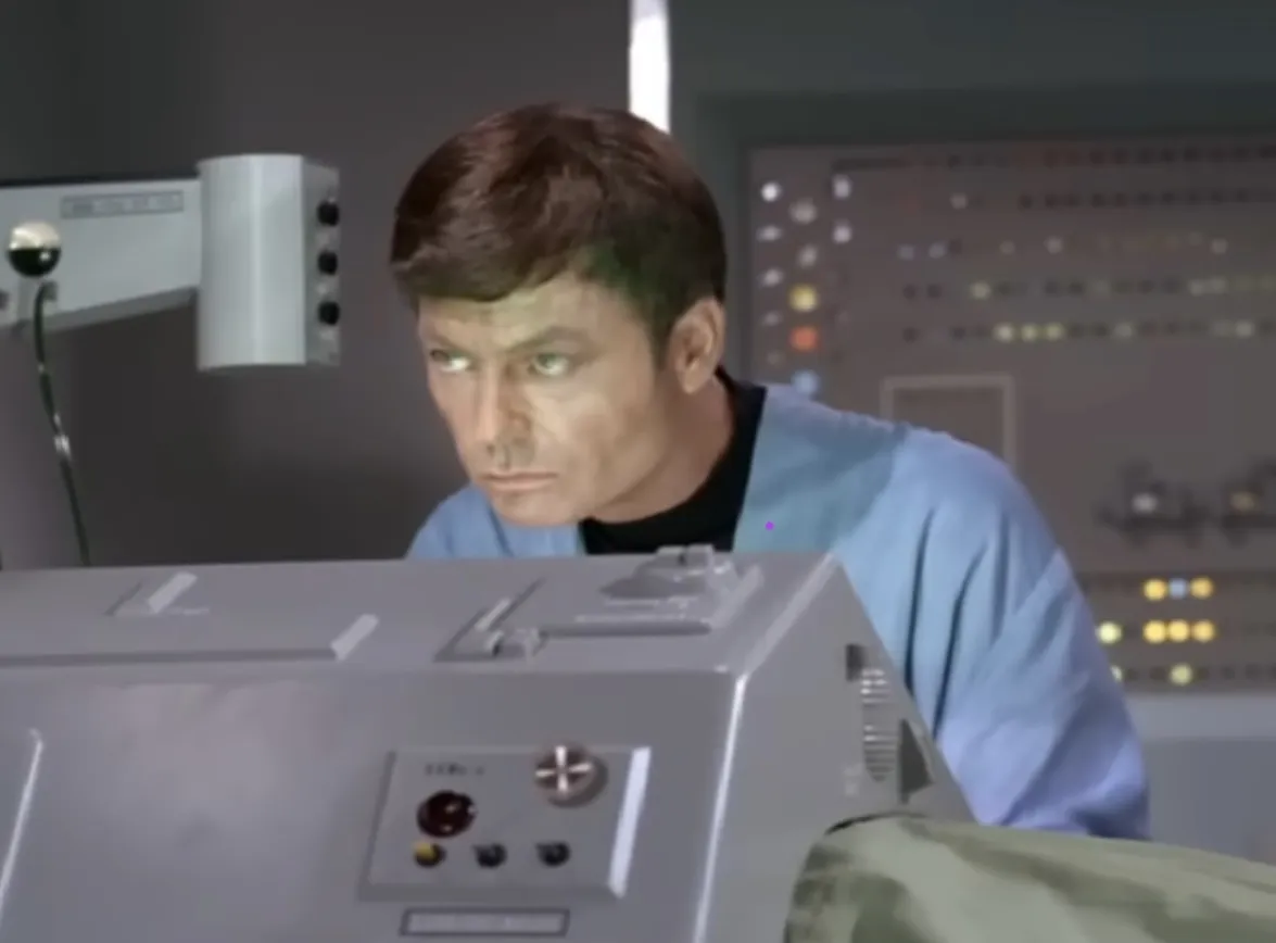 A scene from Star Trek: The Original Series showing Dr. McCoy performing surgery on Sarek. The room is lit with a distinctive green glow. McCoy is centered in the shot, wearing his surgical gown, with retro-futuristic medical equipment surrounding him.