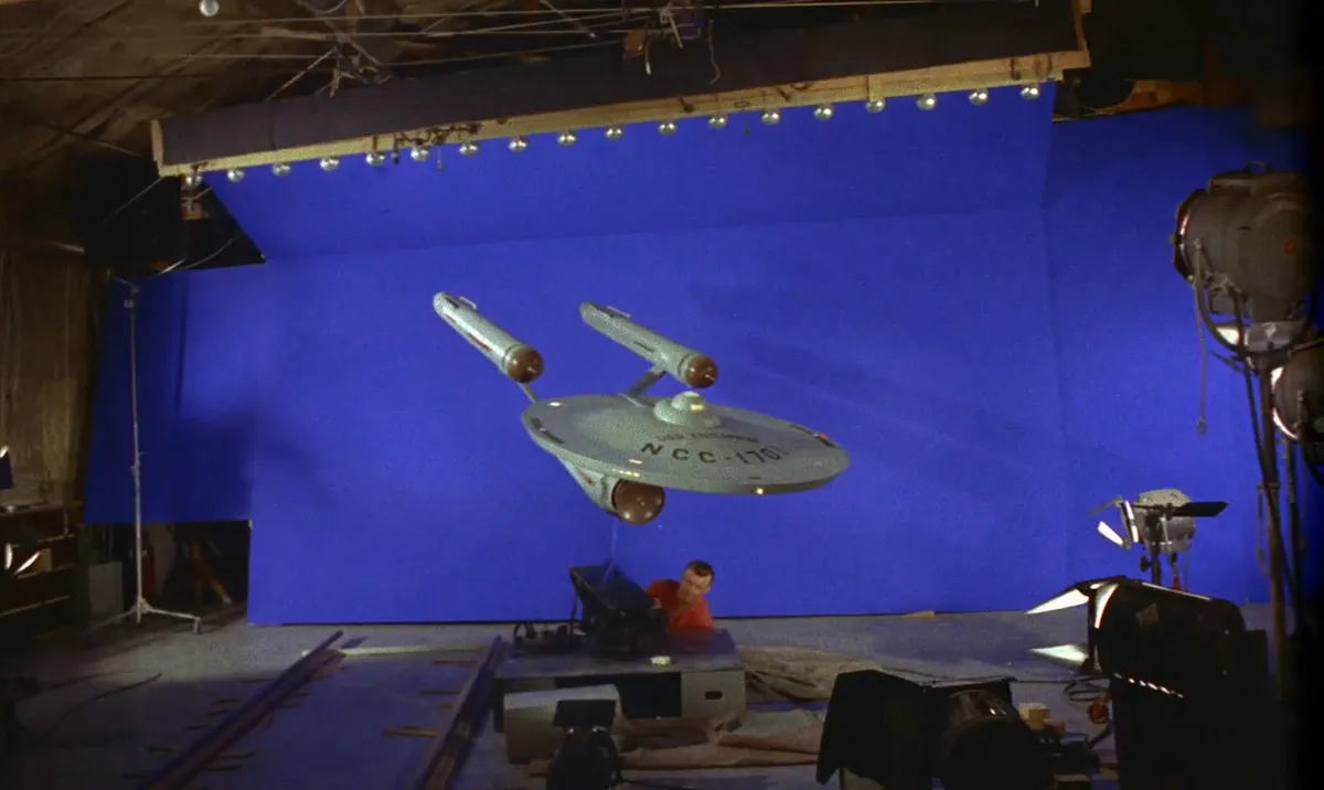 Technicians assemble a model of the USS Enterprise on a Star Trek soundstage, surrounded by lighting rigs. The scene reflects George Merhoff's role as chief gaffer.