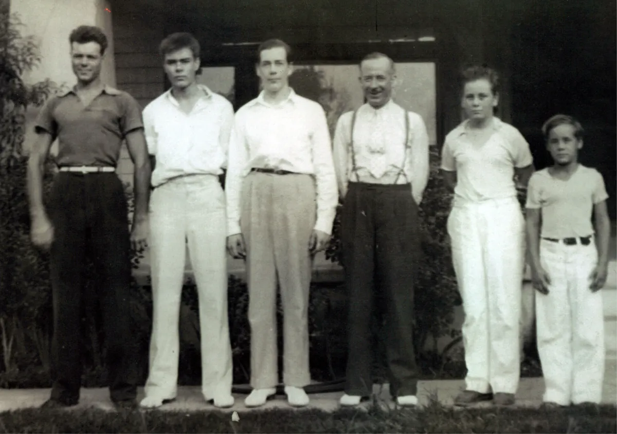 Black and white photo of six Merhoff men standing in a line outside a house. From left to right: George Merhoff (young adult with confident stance), Gene Merhoff (serious expression), Don Merhoff (taller, neatly dressed), Frank Merhoff (older man in suspenders), Paul Merhoff (shorter, youthful face), and Hugh Merhoff (the youngest, standing at the far right).