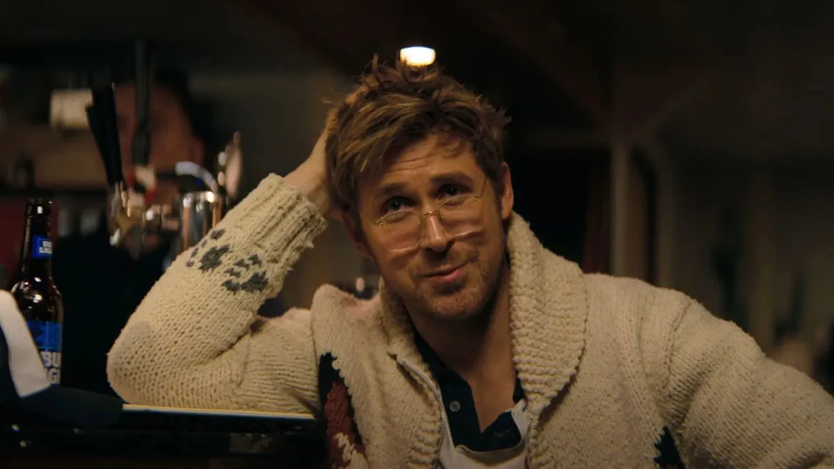 Retrato do ator Ryan Gosling em um ambiente de bar com iluminação amarelada e aconchegante. Ele usa óculos de grau com armação fina, uma camisa polo com detalhes escuros no colarinho e um casaco de tricô grosso na cor creme com padrões geométricos nas mangas. Apoiado sobre o balcão, ele tem a mão direita levantada, tocando os cabelos, e faz uma expressão descontraída com um leve sorriso. Ao fundo, desfocada, é possível ver uma garrafa de cerveja sobre o balcão e silhuetas de outros elementos do bar.