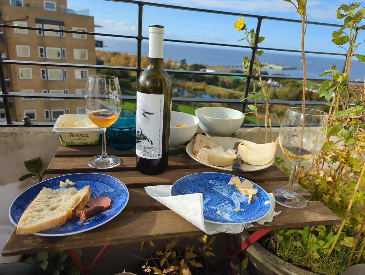 A balcony table set with wine, glasses, bread, and various dishes overlooks a scenic view of a coastal area.