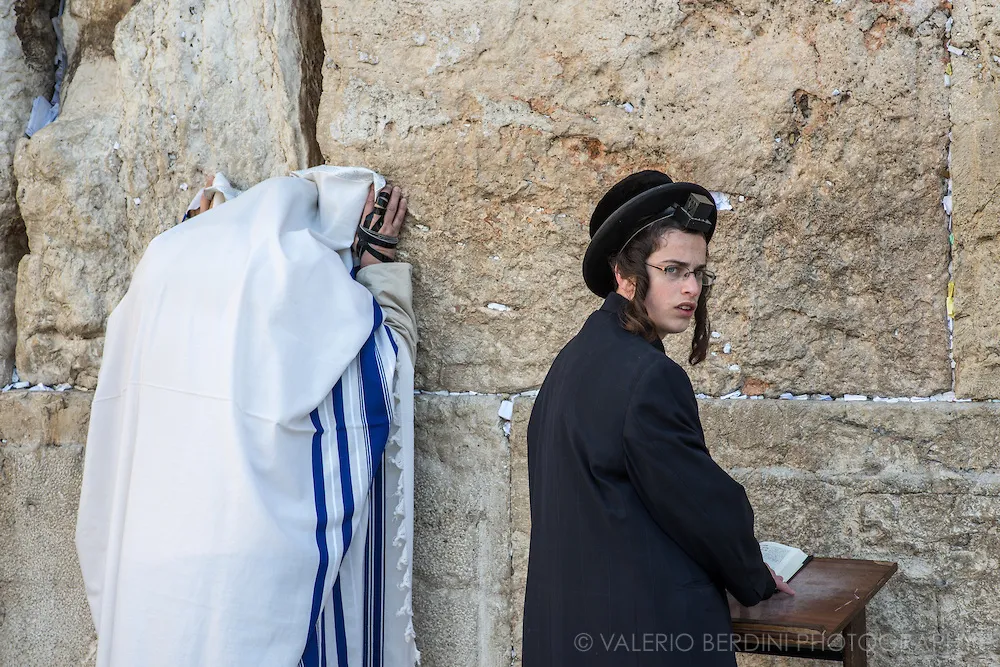 上图：哭墙晨祷。左边的男人披着祷告巾（Tallit Shawl），右手缠着经文匣（Arm Tefillin）。右边的男孩前额头上戴着经文匣(Head Tefillin）。经文匣里面藏着「示马」经文（申六4-9；十一13-21）。