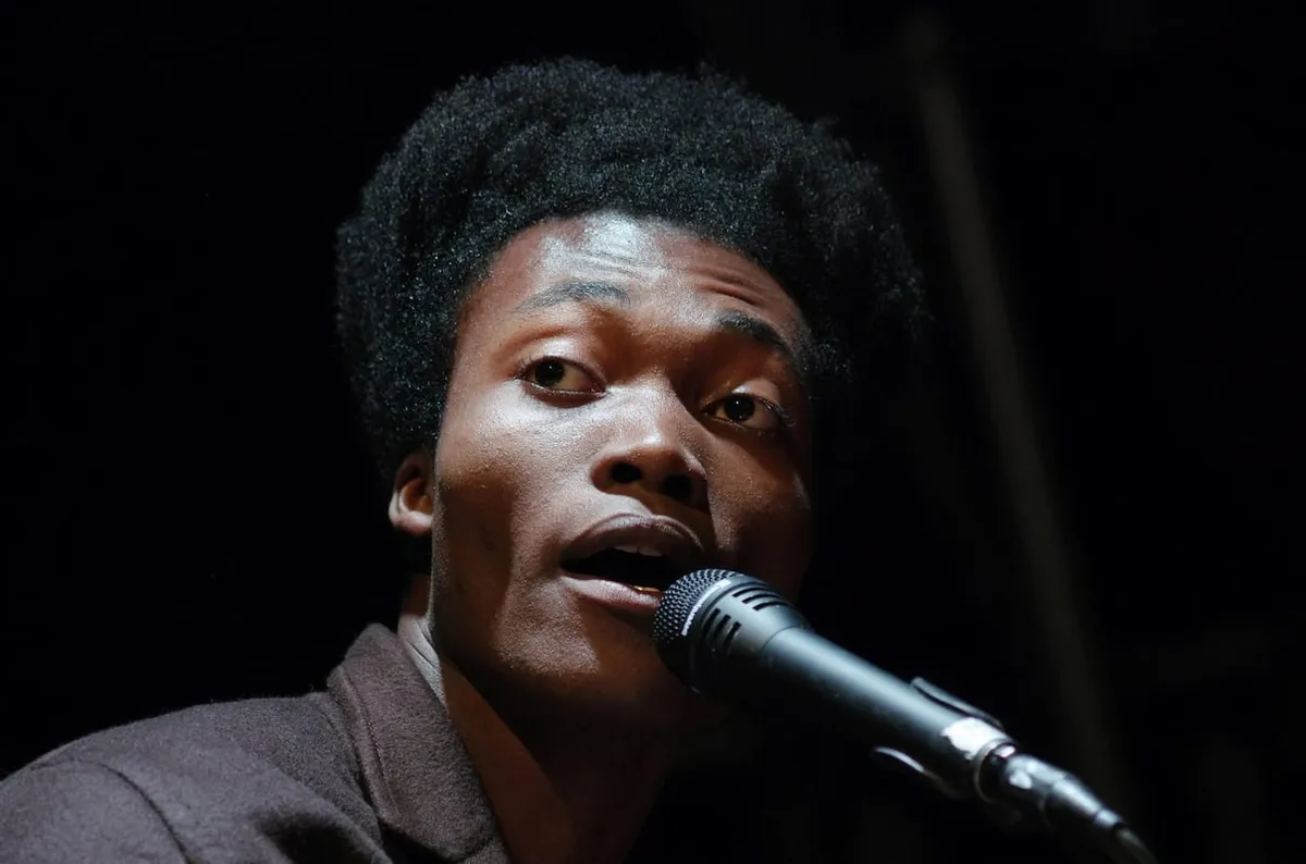 benjamin clementine singing into a black microphone in front of a black background.