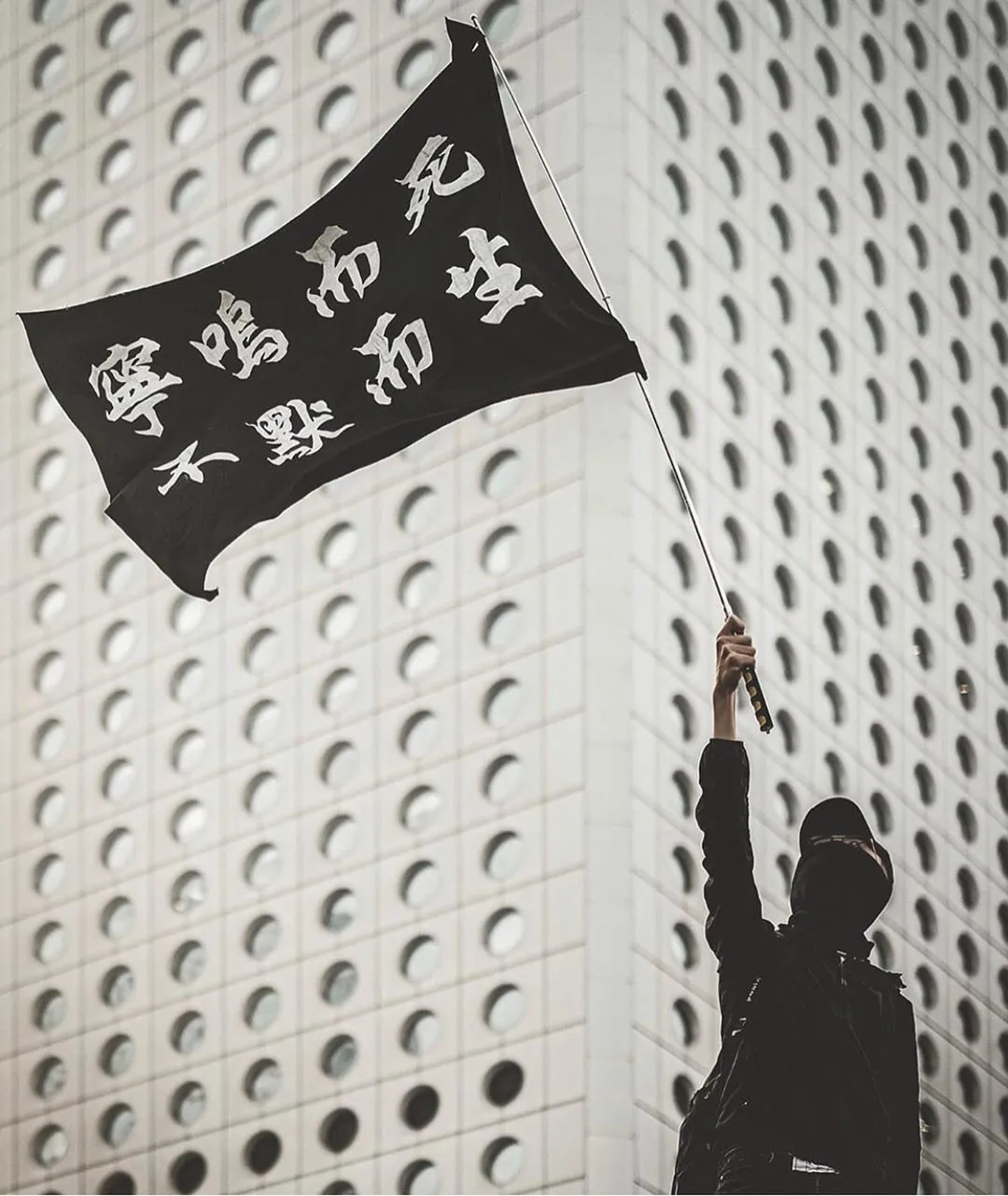 A masked protester in black raising a black flag reading 寧鳴而死，不默而生