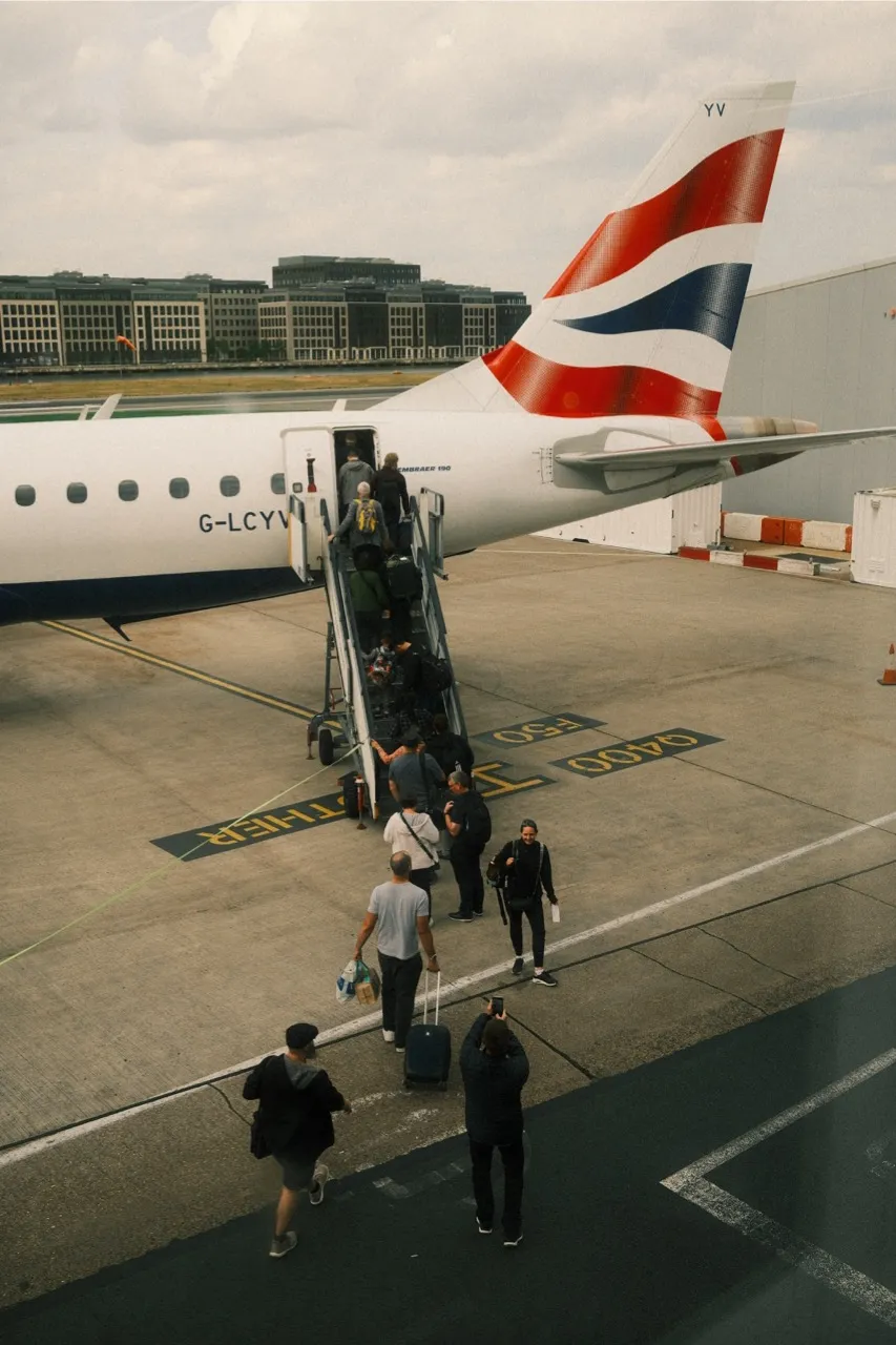 Passengers boarding a British Airways aircraft via mobile stairs on an airport tarmac. The white aircraft displays the distinctive red, white, and blue Union Jack livery on its tail. Ground crew and passengers with luggage are visible around the aircraft, with modern airport buildings in the background under an overcast sky.