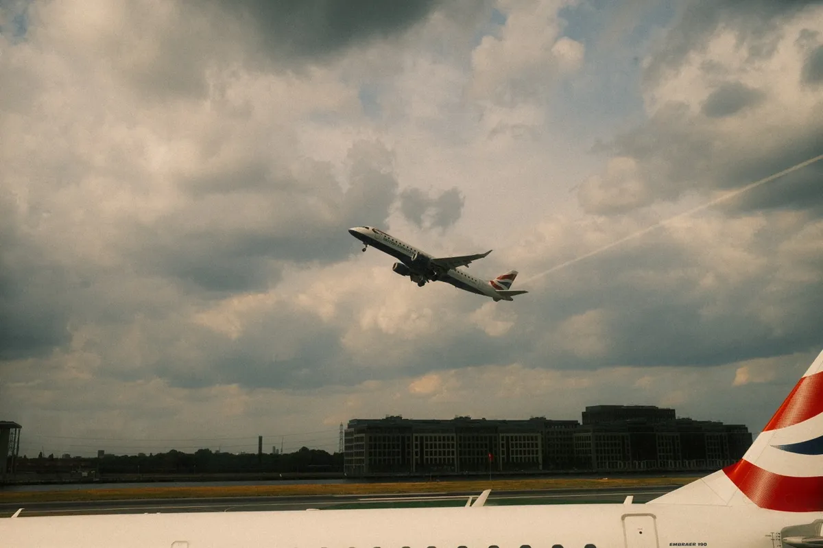 A British Airways aircraft taking off against a dramatic cloudy sky. The aircraft is captured mid-flight with airport buildings visible in the background. Part of another aircraft's wing with British Airways livery is visible in the foreground.