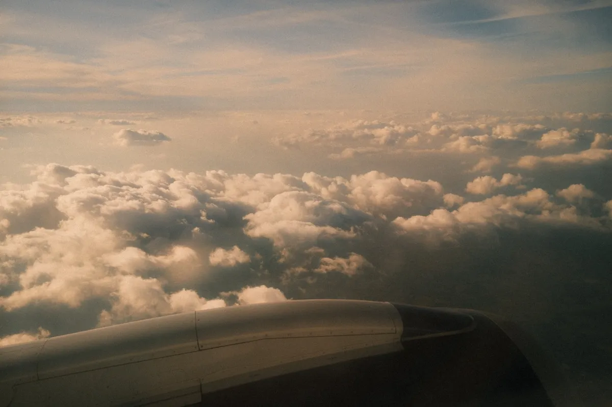 Aerial view from an aircraft window showing a dramatic cloudscape below. Thick, billowing white and gray clouds stretch across the frame, with the aircraft's white wing visible in the lower portion of the image. The lighting suggests either sunrise or sunset conditions.