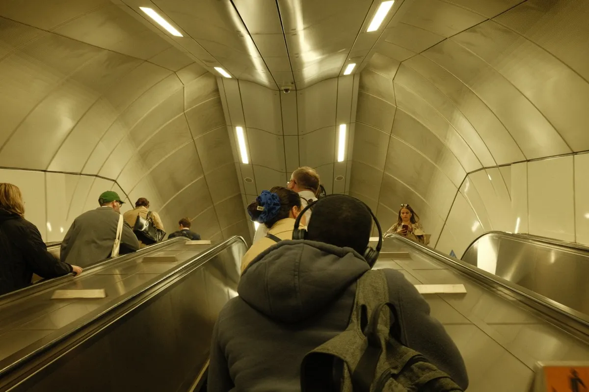 Interior view of a London Underground escalator tunnel showing passengers descending. The characteristic curved white-tiled ceiling with fluorescent lighting creates the iconic tube tunnel aesthetic. Several people are visible on the escalator, with one person prominently featured in dark clothing.