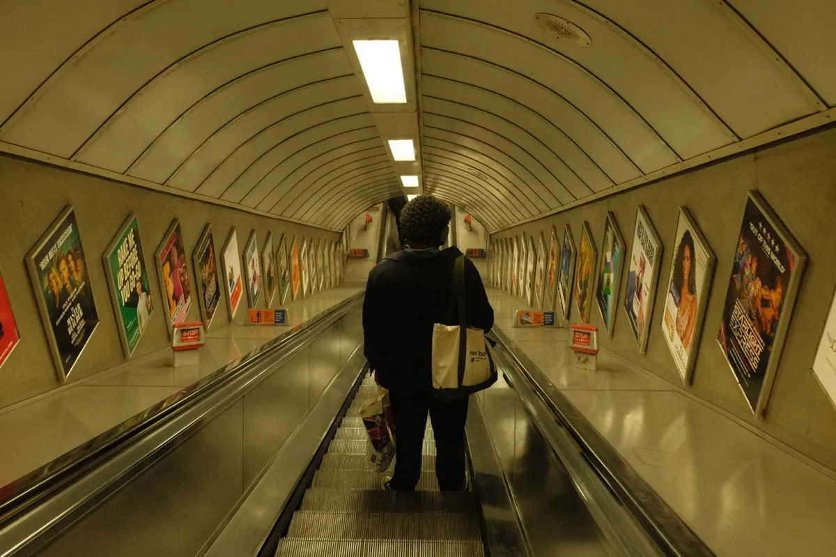 A person walking down a London Underground escalator in a tunnel lined with advertising posters. The curved tunnel features the typical white tiled walls with colorful advertisements on both sides. The perspective shows the length of the escalator descending into the station.
