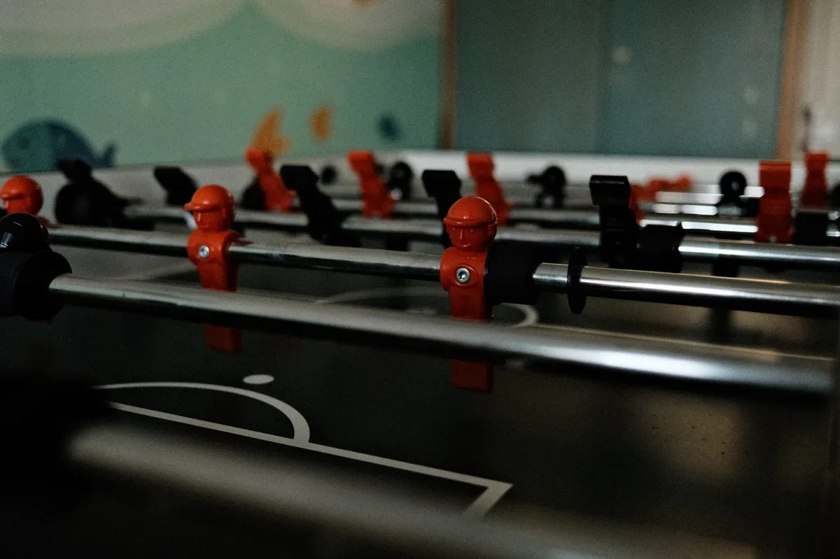 Foosball players, positioned on metal rods, stand ready on a table. The scene is set against a blurred, colorful wall that suggests a playful environment.