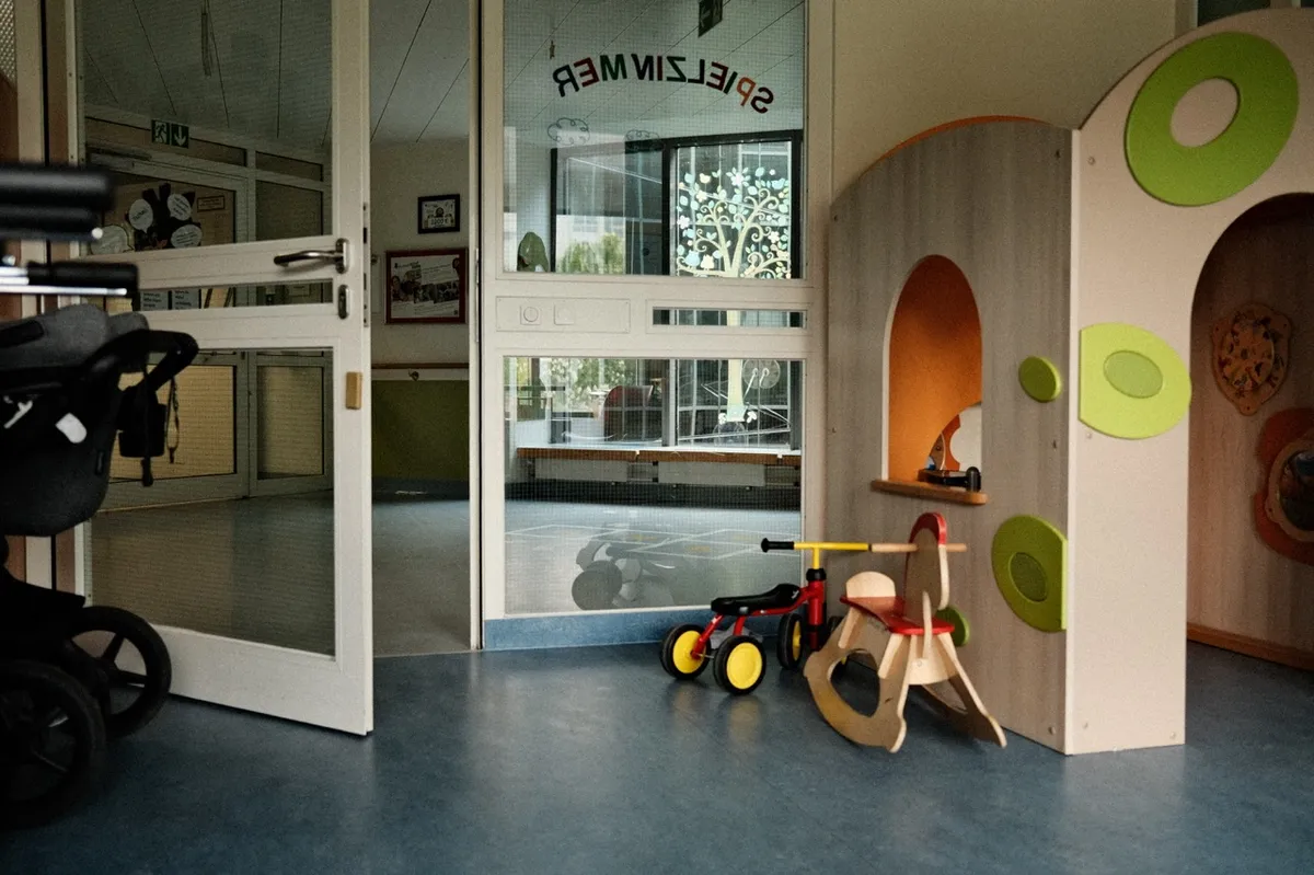 Children’s playroom with a tricycle and rocking horse set against colorful play structures. A stroller rests by an open door. “SPIELZIMMER” is written on a glass panel.