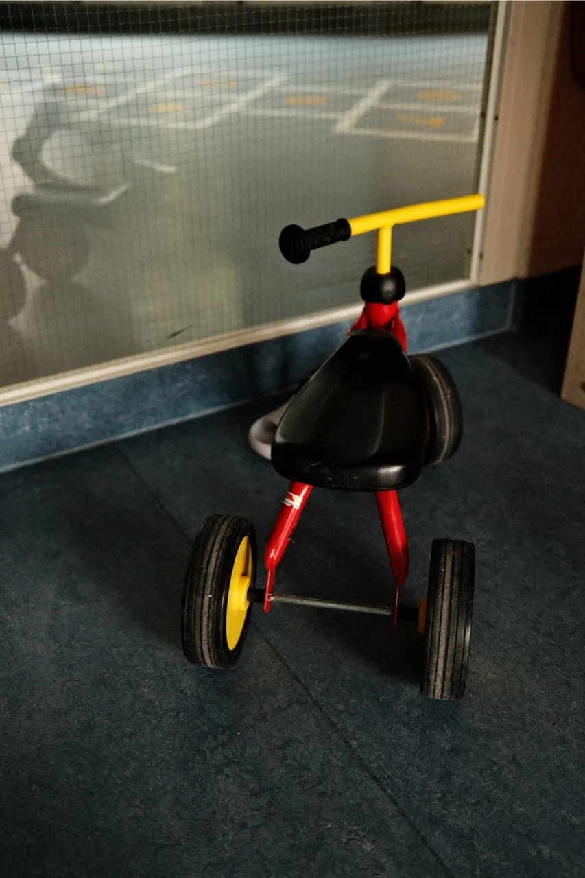 A small tricycle with yellow and black wheels is parked on a blue tiled floor, in front of a partially transparent glass door.