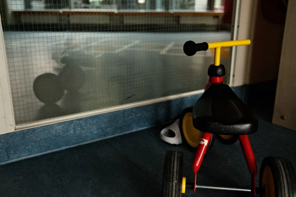 Red and yellow tricycle is parked indoors beside a mesh glass panel, casting a shadow. Nearby, a pair of white sandals rests on the blue-tiled floor.