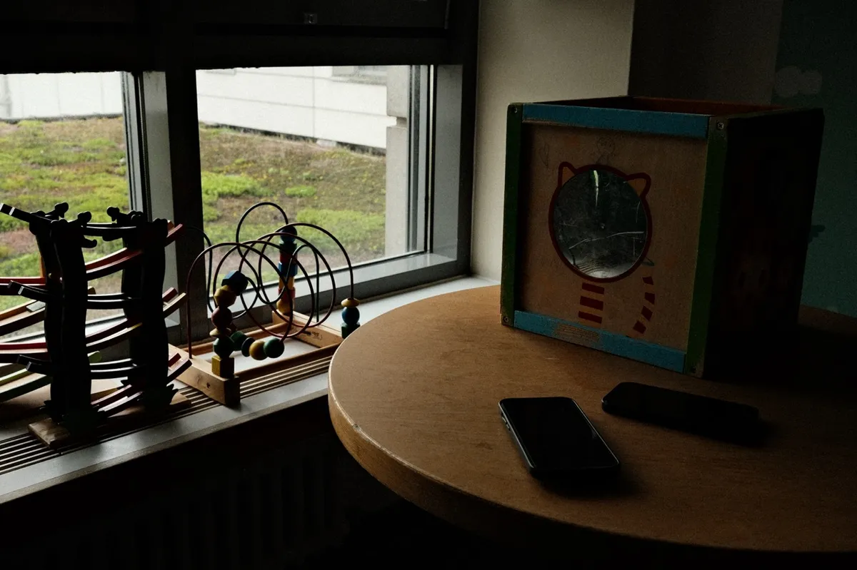Toys rest on a window sill, including a bead maze and a wooden box with a cat design. A phone lies on a round table, with a view of greenery outside.