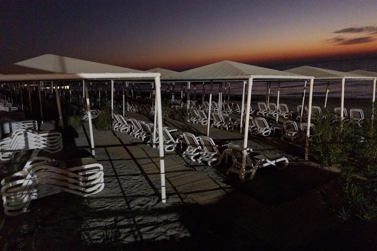 Lounge chairs are stacked under white canopies, resting on a wooden path along a dimly lit, deserted beach at twilight, with an orange glow on the horizon.