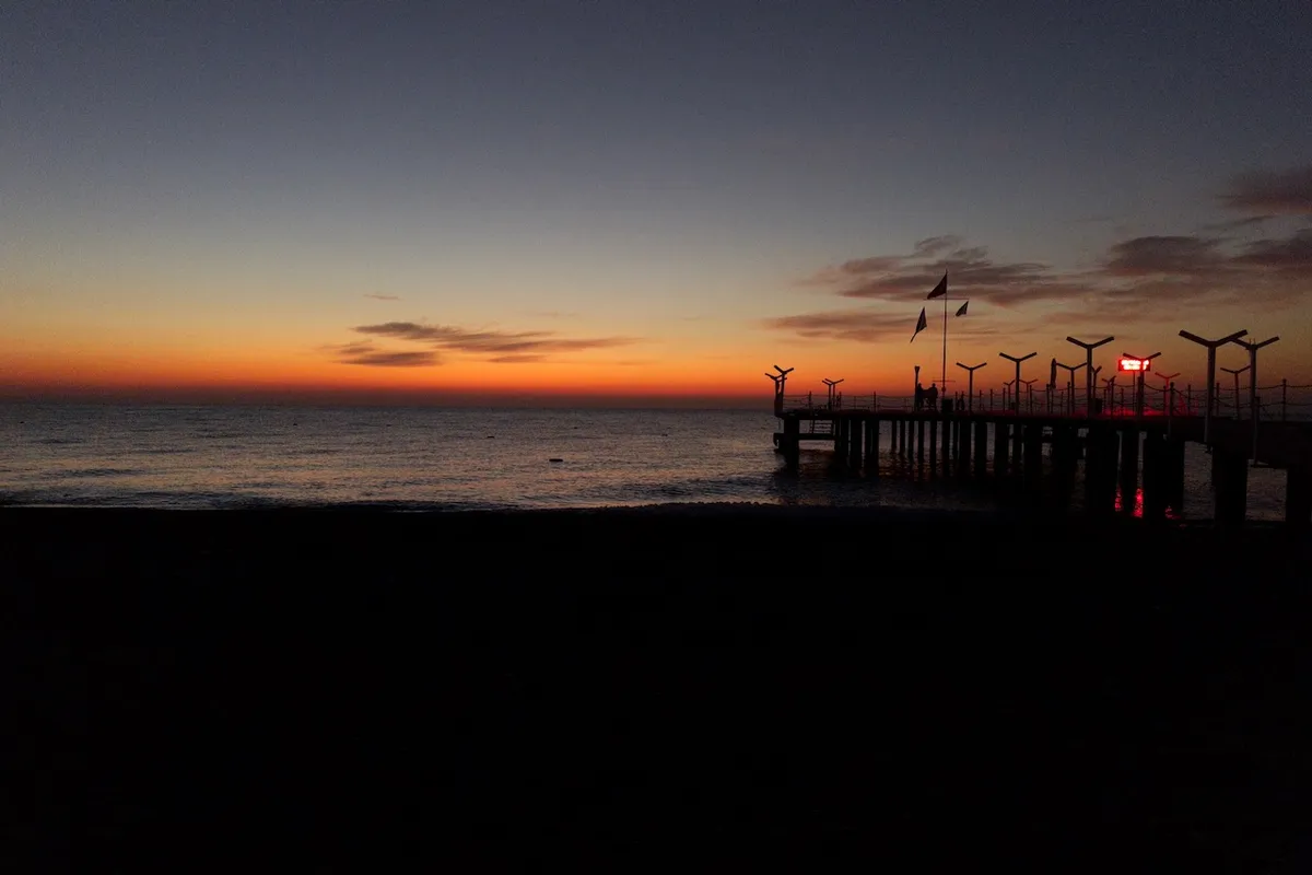Pier extends over calm sea at sunrise, silhouetted against a gradient sky of deep orange to dark blue, with scattered clouds and distant flags fluttering.