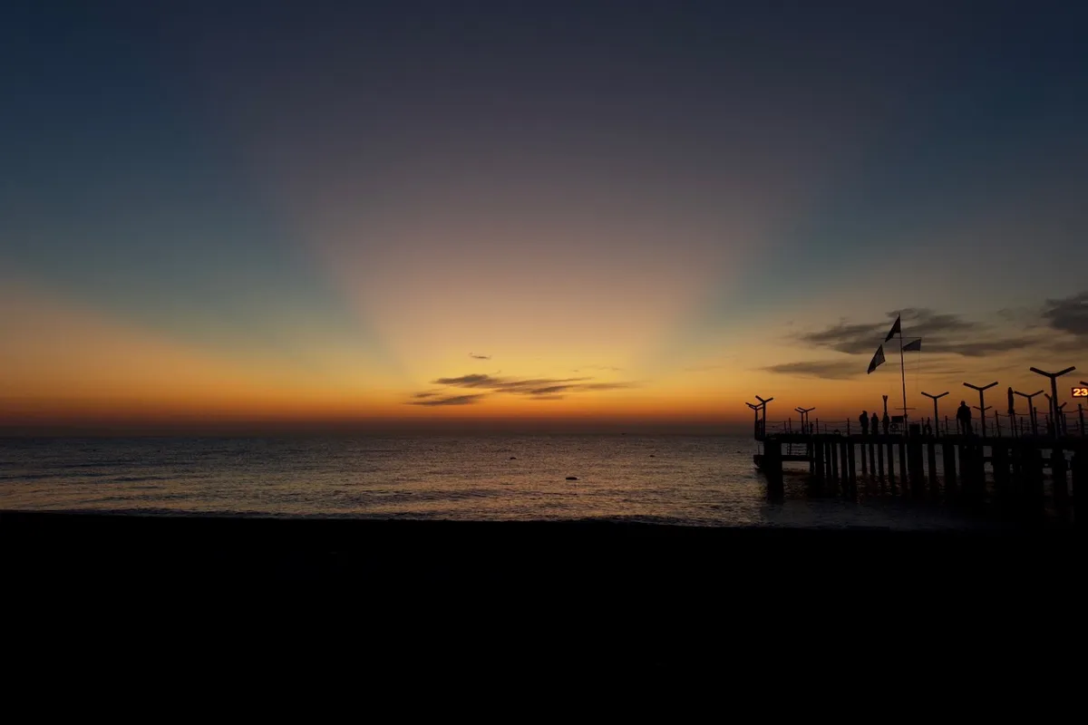 Sunrise casts vibrant hues over a calm ocean, silhouettes of figures and flags stand on a pier extending into the water, enhancing the tranquil scene.