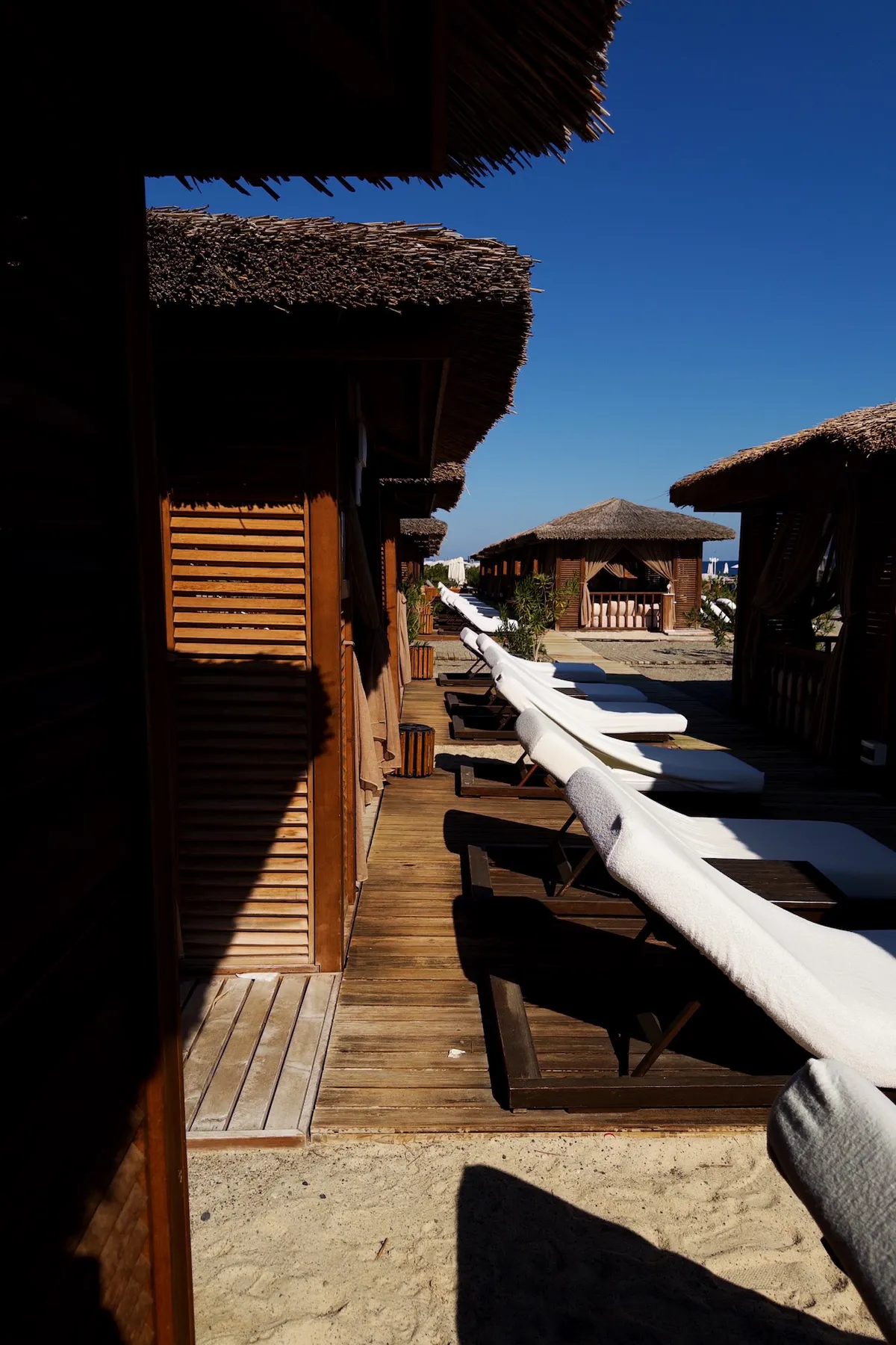 Wooden cabanas line a sandy beach, with white cushioned lounge chairs stretching along wooden decks under a clear blue sky, creating a serene and tropical resort atmosphere.