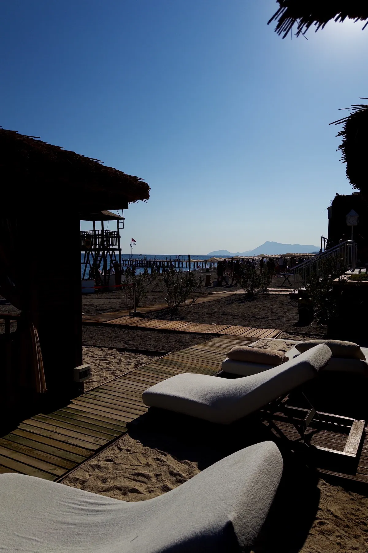 Lounge chairs rest on a wooden deck, facing a sandy beach with people walking near a pier. Mountains and a clear blue sky provide a serene backdrop.