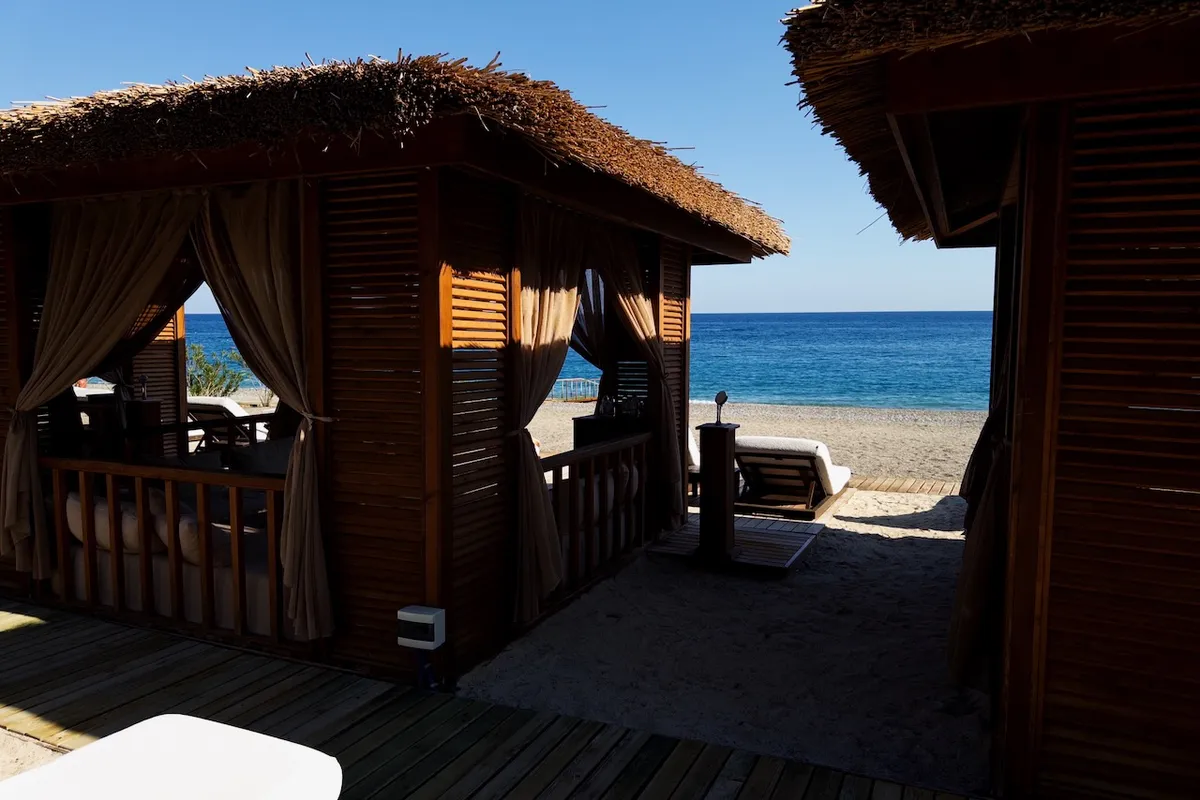 Thatched cabanas stand beside the ocean, with wooden slats and curtains offering shade. The sandy beach stretches to the blue sea under a clear sky.