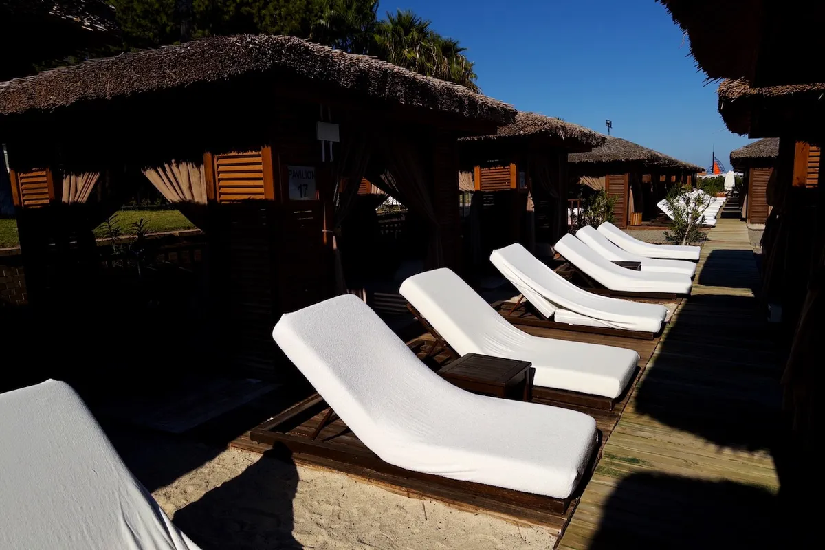 White lounge chairs are arranged in a row on a wooden deck, shaded by thatched cabanas. In the background, lush greenery and a clear blue sky enhance the serene atmosphere. A sign reads "PAVILION 17."