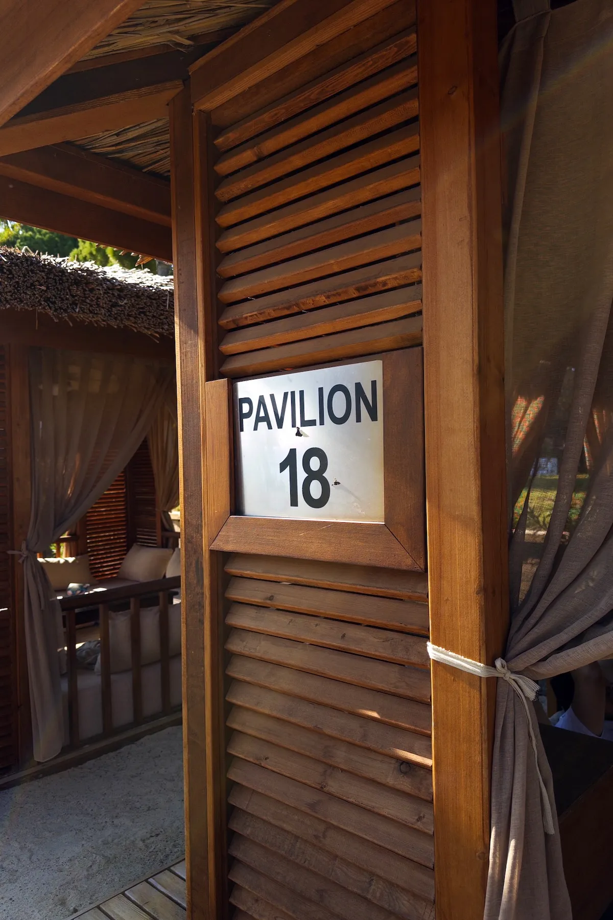 A wooden pavilion displays a sign labeled "PAVILION 18." The structure features slatted walls and tied-back curtains, situated in a sunlit outdoor setting.