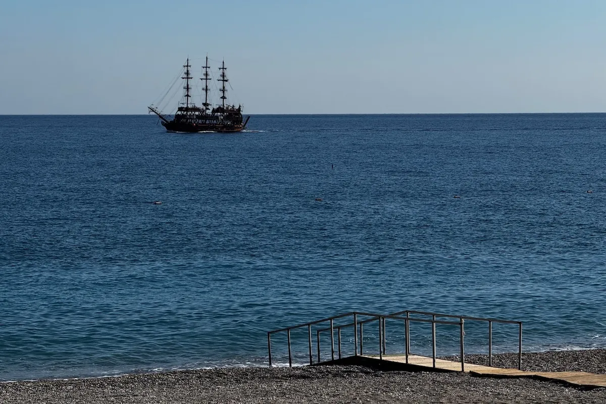 A tall ship with three masts sails on calm blue ocean waters, while a wooden ramp extends toward a pebble beach in the foreground under a clear sky.