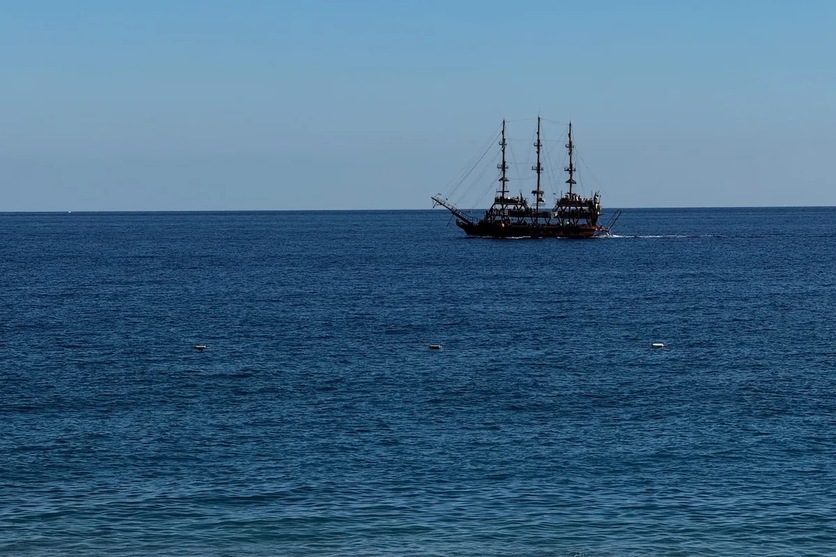 A sailing ship with multiple masts moves across the calm sea, under a clear blue sky, suggesting a peaceful maritime setting.