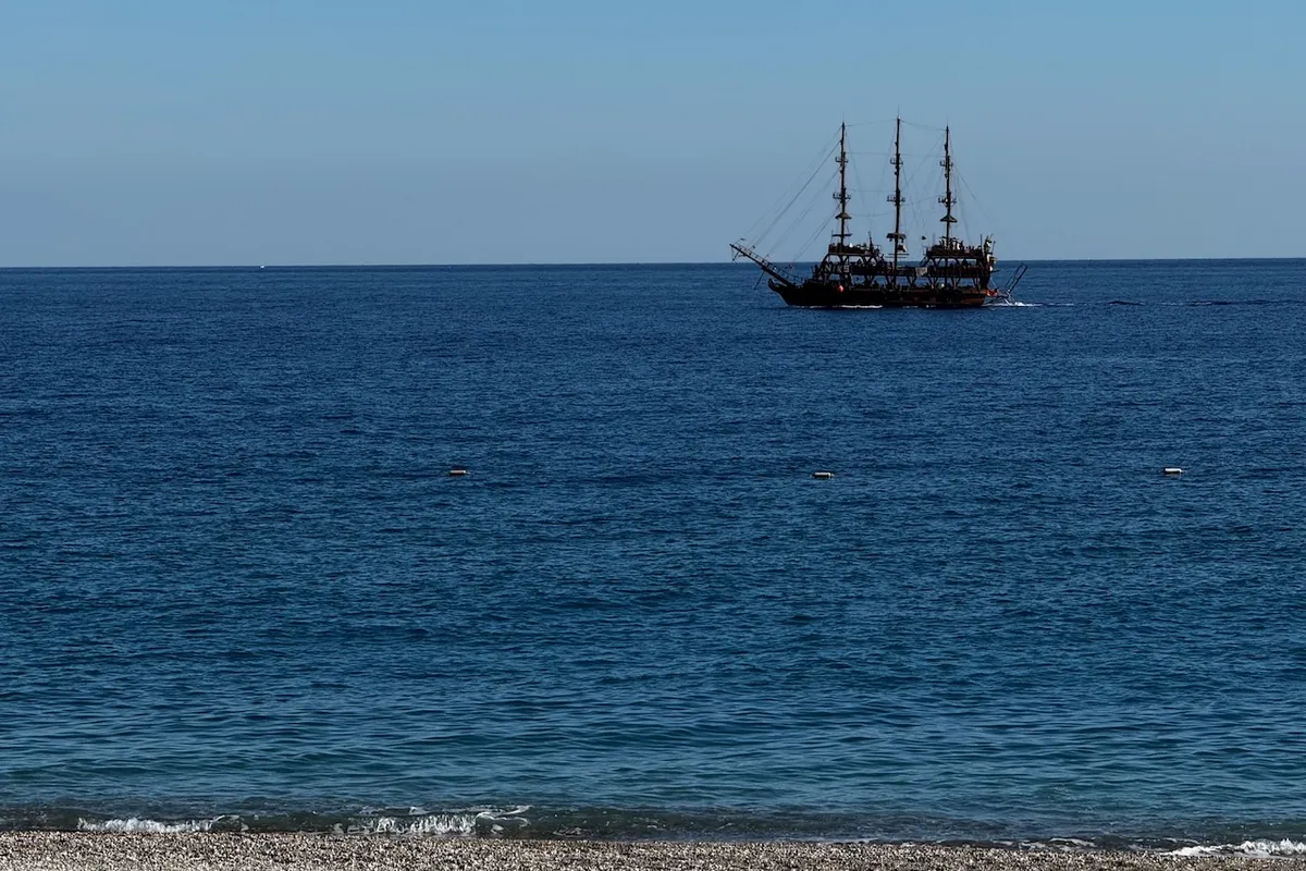 A large sailing ship with multiple masts sails on a clear blue sea, near the horizon, under a cloudless sky, with a pebbled beach in the foreground.