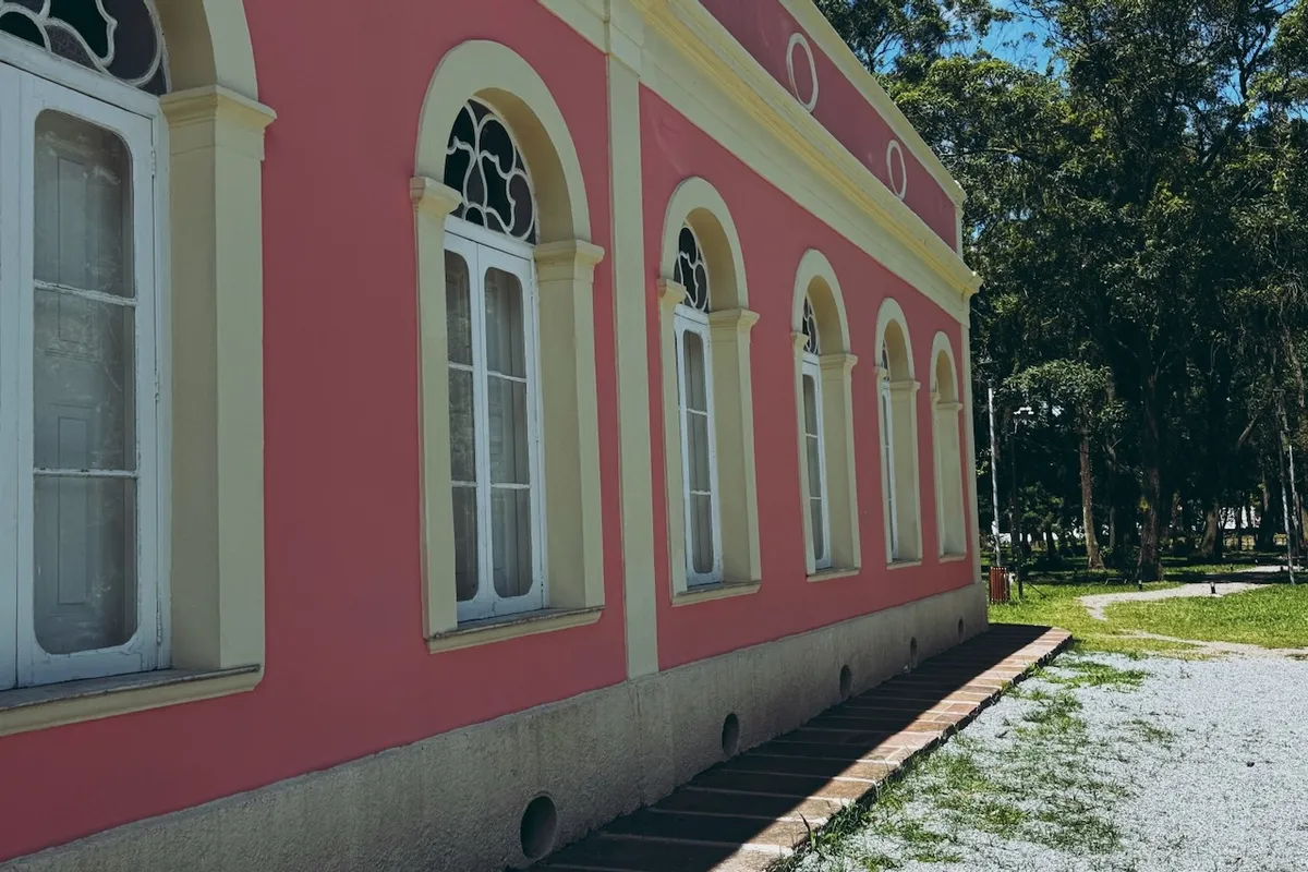 A pink building with white arched windows stands in the sunlight beside a gravel path. Surrounding are green trees and a grassy area, indicating a park or garden setting.