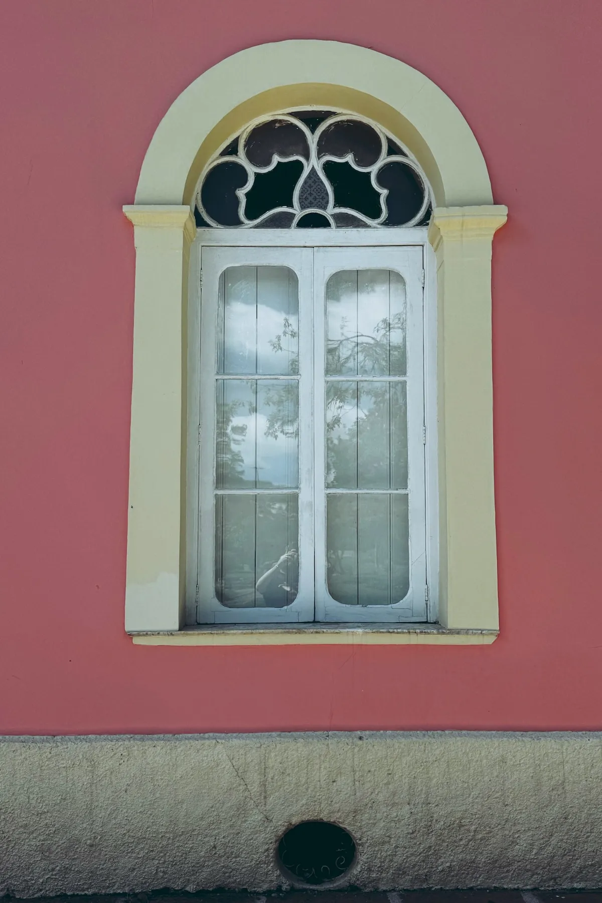 An arched window with decorative glass panes reflects trees, set in a pink wall and bordered by a light yellow frame, above a textured beige base.