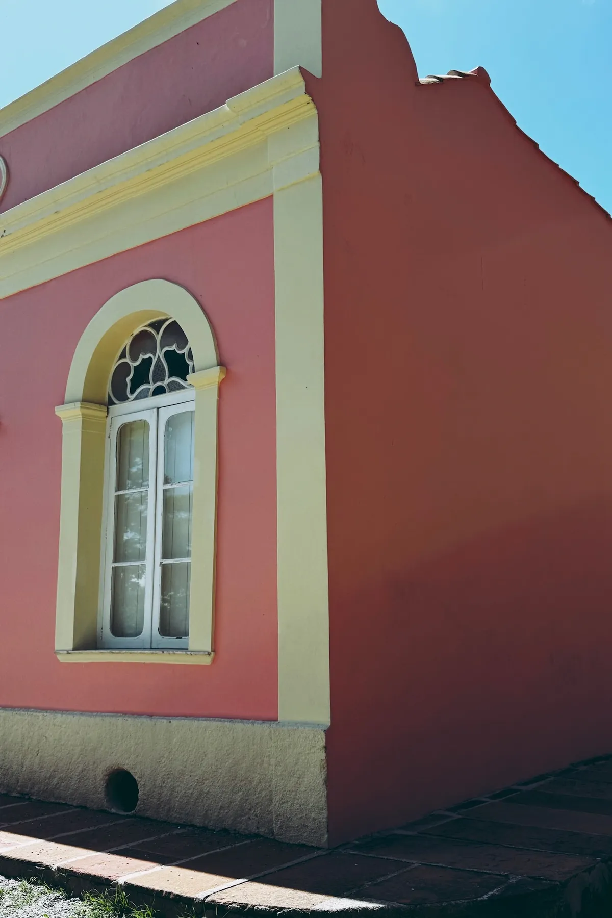 A pastel pink and yellow building stands under the clear blue sky, featuring an arched window with intricate detailing, set on a stone pavement.