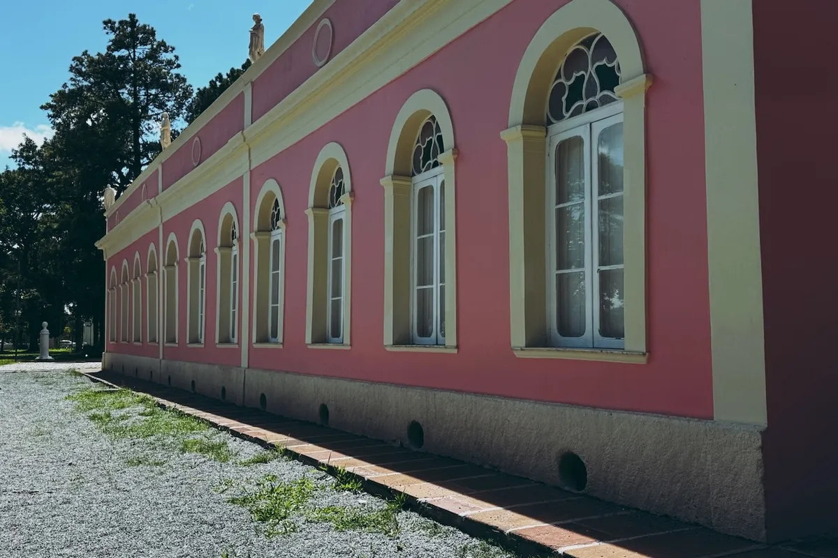 A pink, classical-style building with arched windows sits along a gravel path. Sculptures adorn the roof, surrounded by trees and a clear sky.