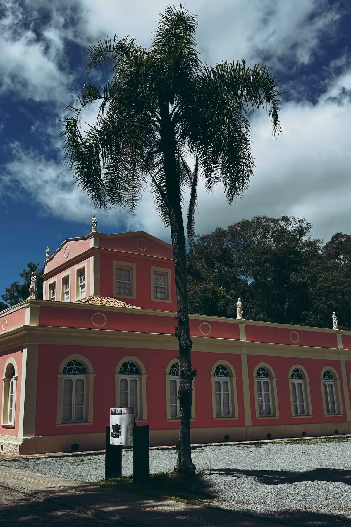 A tall palm tree stands in front of a pink building with arched windows and statues on the roof, set against a partly cloudy sky and surrounded by greenery.