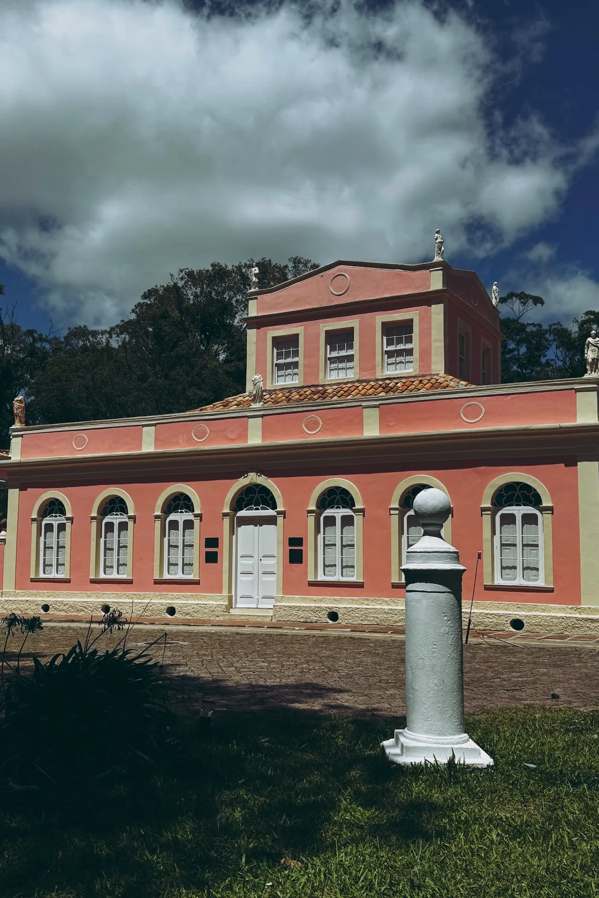 Pink and cream neoclassical building stands with arched windows, surrounded by trees and topped with statues. A white pillar is in the foreground on a grassy lawn under a cloudy sky.