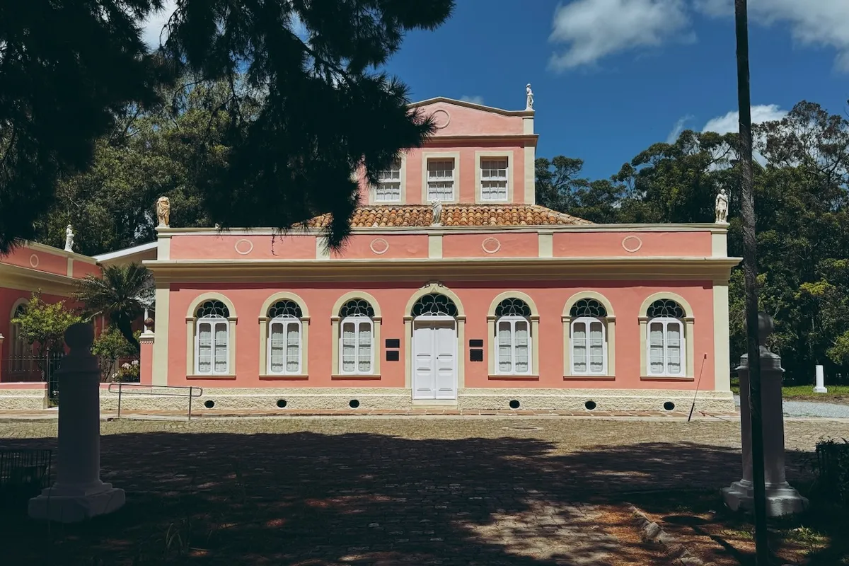 A pink, single-story building with a white door and arched windows stands in a sunlit courtyard, surrounded by trees and blue sky. Statues are positioned atop the roof.