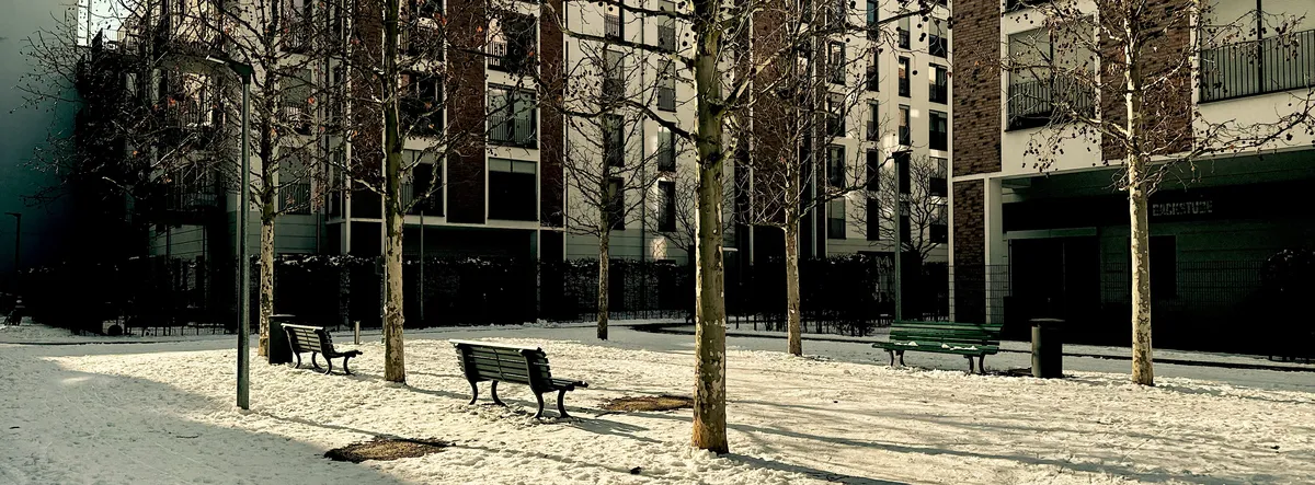 Benches sit empty in a snowy courtyard, surrounded by leafless trees. Modern apartment buildings form the urban backdrop in low sunlight.