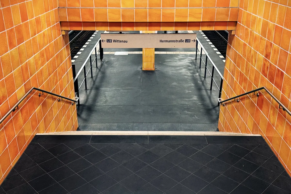 A view of a subway station entrance with stairs leading down to the platform. The walls are covered in bright orange tiles. A sign overhead indicates directions for the U8 line, with arrows pointing left to Wittenau and right to Hermannstraße. The platform area is visible in the background, with black railings and railway tracks.