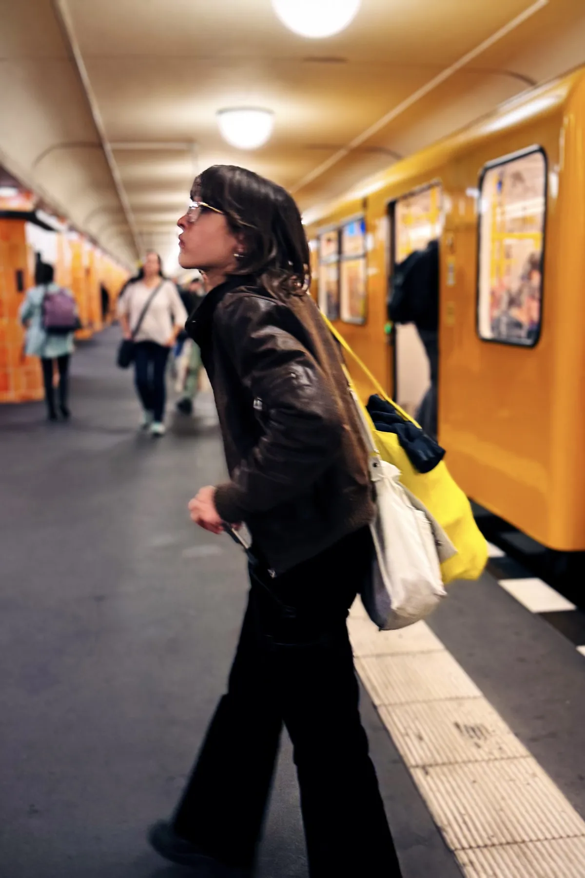 A person in a brown jacket and glasses is looking down the platform in a subway station. They are holding a white and yellow bag. A yellow train is stopped at the platform, and people can be seen walking in the background. The station has a curved, well-lit ceiling.