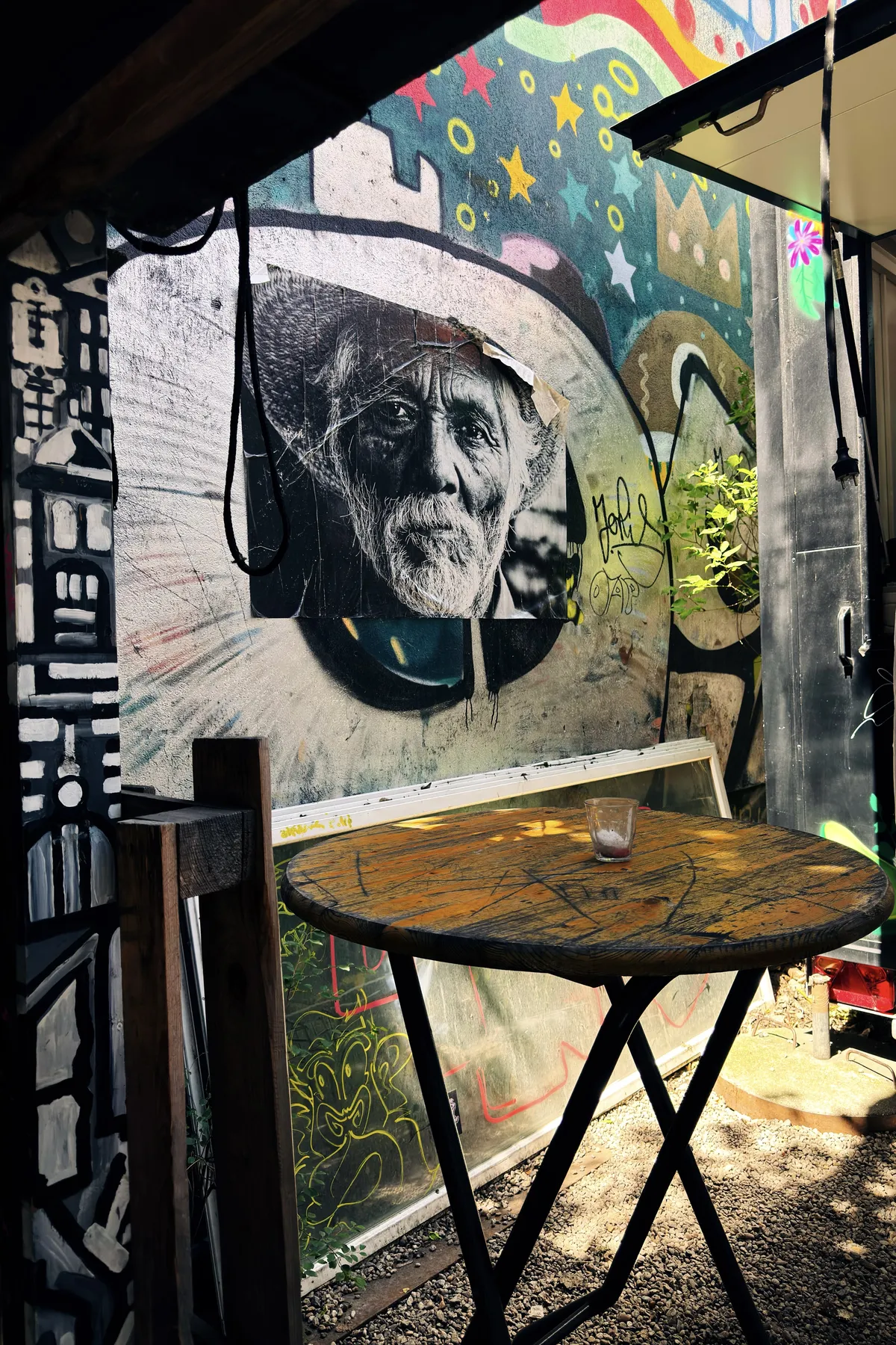 An outdoor scene featuring a wooden table with a glass placed on it. The surrounding walls are covered in colorful graffiti, including stars and abstract designs. A black and white poster of an elderly man is displayed on one wall, adding a striking contrast to the vibrant artwork. Sunlight filters through, casting shadows on the gravel floor.