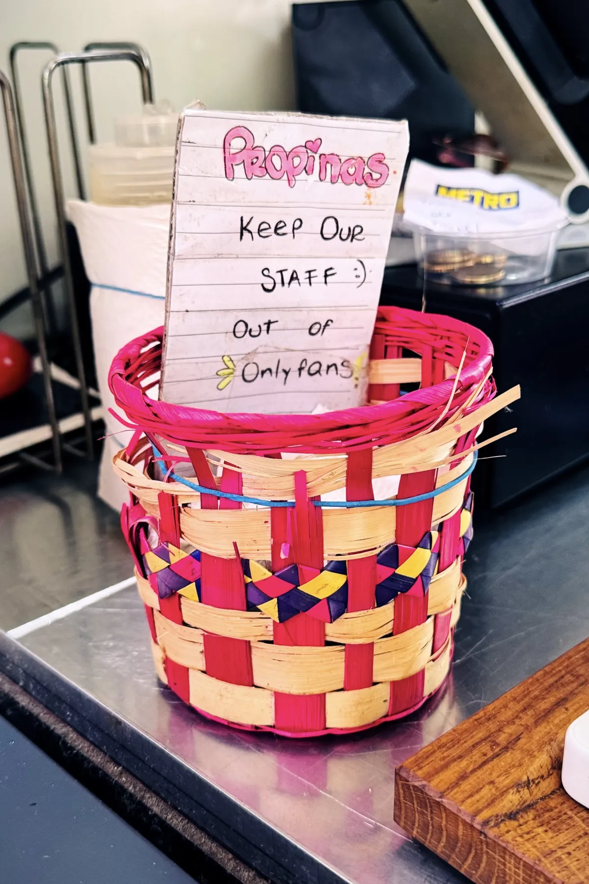A colorful woven basket sits on a counter with a humorous handwritten sign inside. The sign reads "Propinas: Keep Our Staff :) Out of OnlyFans" in playful lettering. The background shows part of a restaurant workspace, with a holder for napkins and some coins in a small plastic container.
