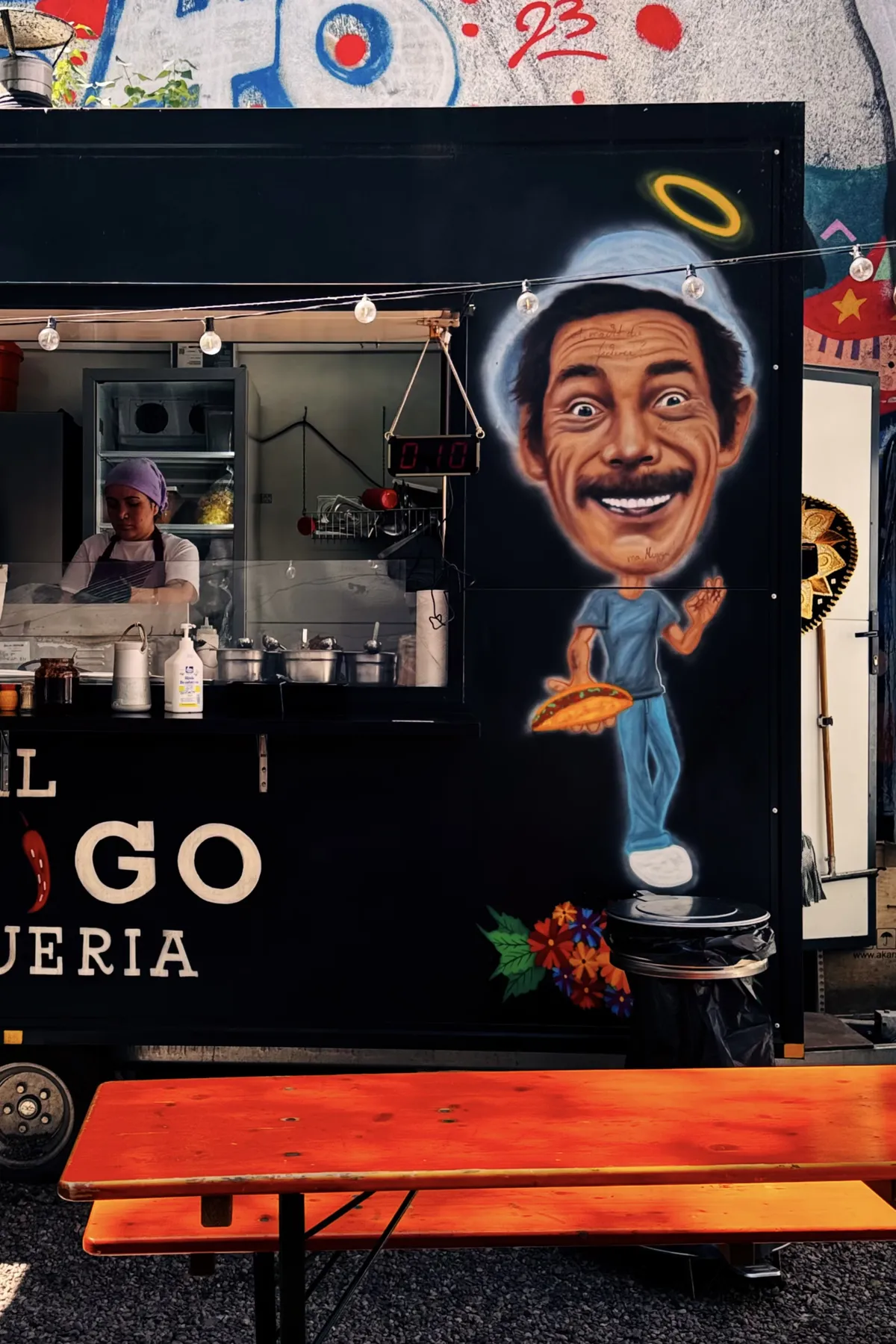 A vibrant food truck with a mural of a smiling man holding a taco, adorned with a halo. The truck features a counter with various condiments and a staff member preparing food. Bright red picnic tables are in front of the truck, and there's colorful graffiti art on the surrounding walls.
