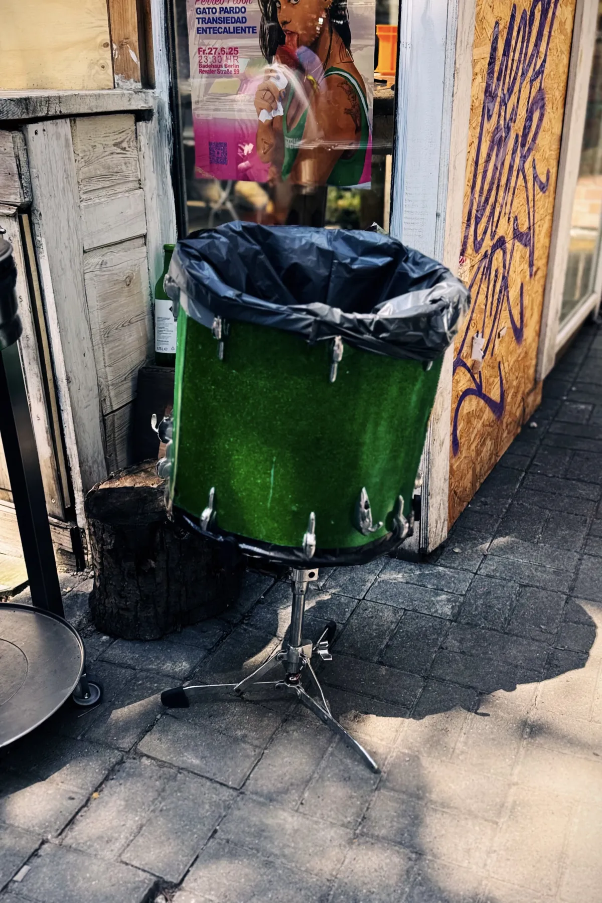 A green bass drum has been repurposed as a trash can and is placed on a sidewalk, with a black trash bag lining its interior. The drum is on a metal stand, next to a wall with graffiti and a poster featuring artwork of a person in a green tank top. A small tree stump is also visible nearby.