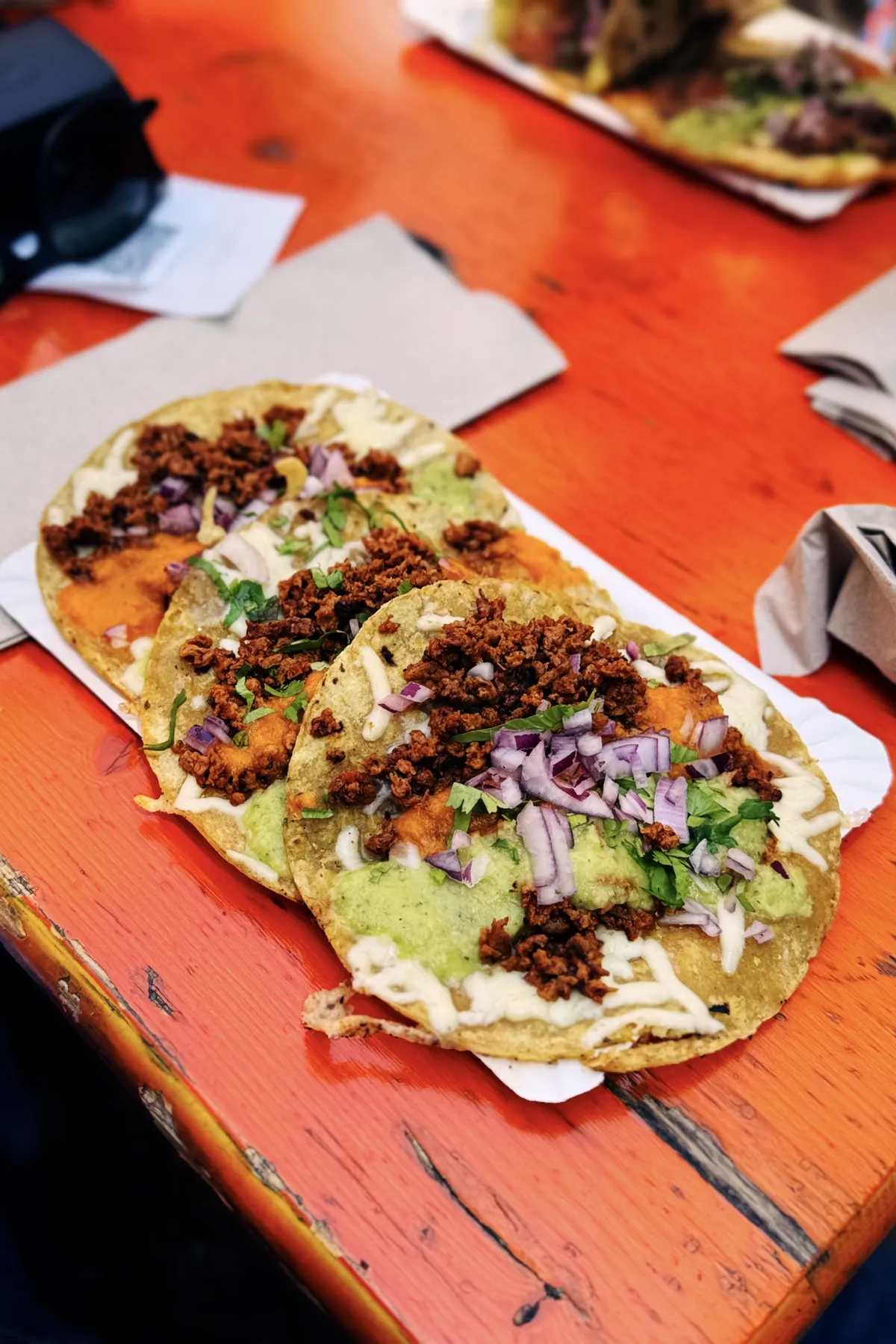 Three tacos are laid out on a white paper tray atop a bright orange wooden table. The tacos are topped with crumbled veggie chorizo, diced red onions, fresh cilantro, and a blend of green and orange sauces. A couple of napkins are visible next to the tacos, and a blurry glimpse of another similar dish is seen in the background.