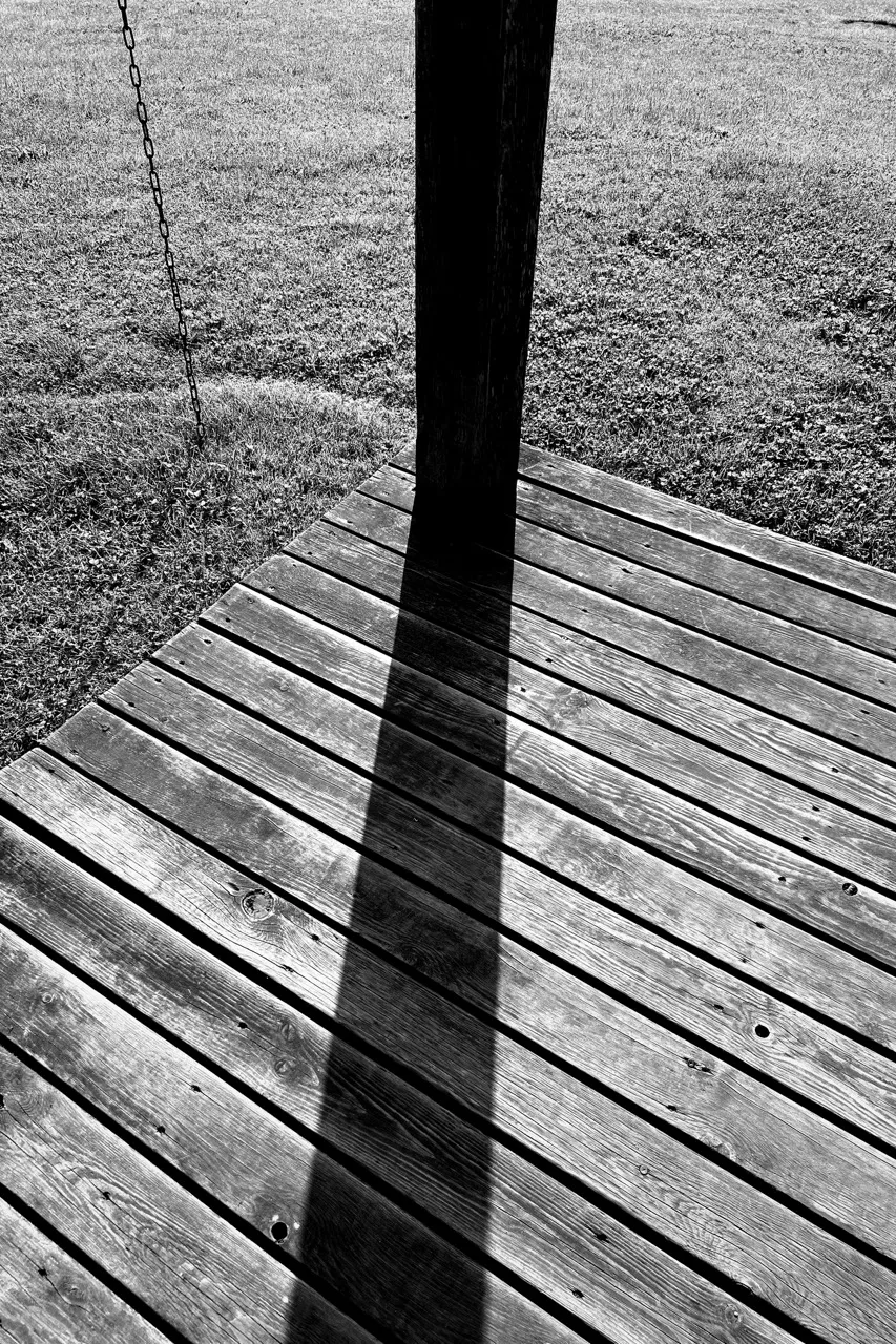 Wooden deck casts a long shadow from a vertical post, while a suspended chain hangs nearby. The deck overlooks a grassy field under bright sunlight.