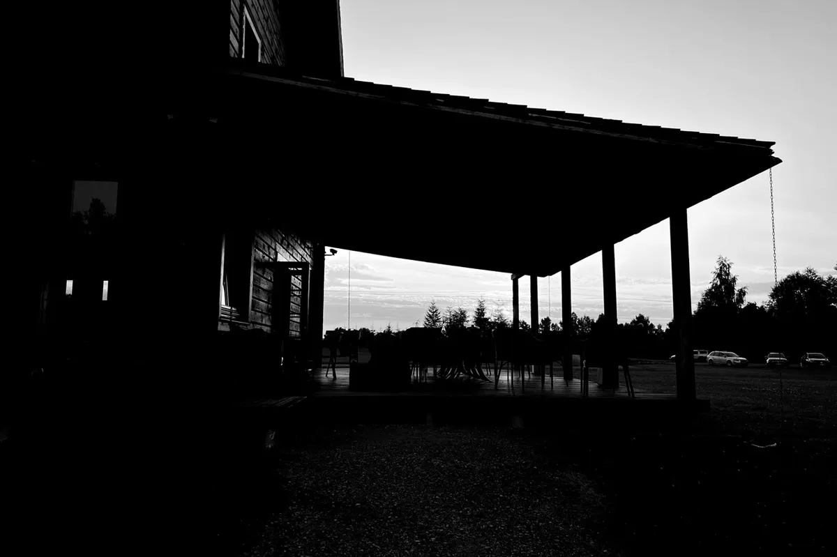 Silhouetted wooden house with a large covered patio, stands against a cloudy sky. Trees and parked cars are visible in the distance, creating a serene, rural setting.