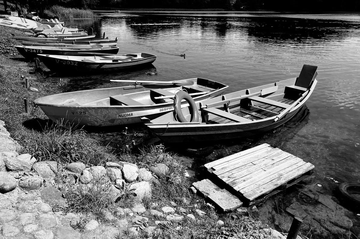 Rowboats rest on a lakeshore, lined up beside wooden docks in calm water. Nearby stones and grass frame the scene. Visible text on a boat includes "NUOMA" and "V-25".