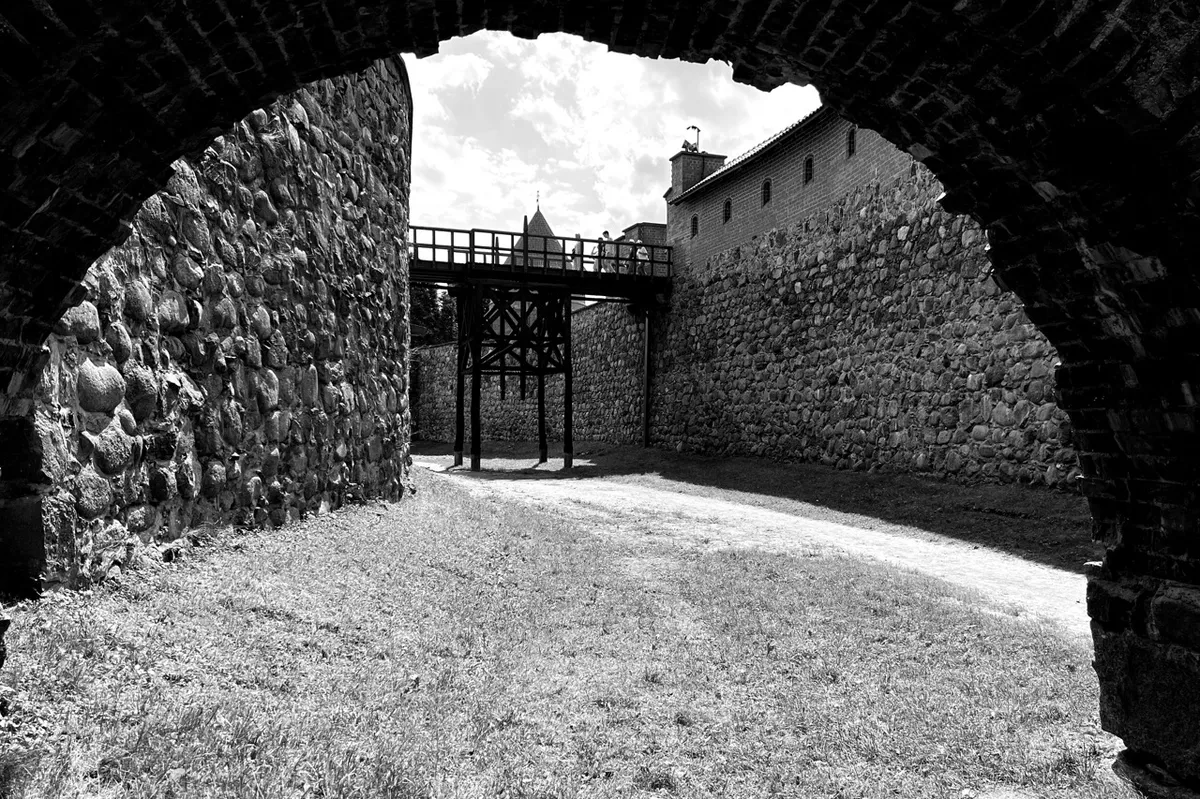 Arched stone entryway frames a view of an elevated wooden bridge between two ancient stone fortress walls under a cloudy sky.