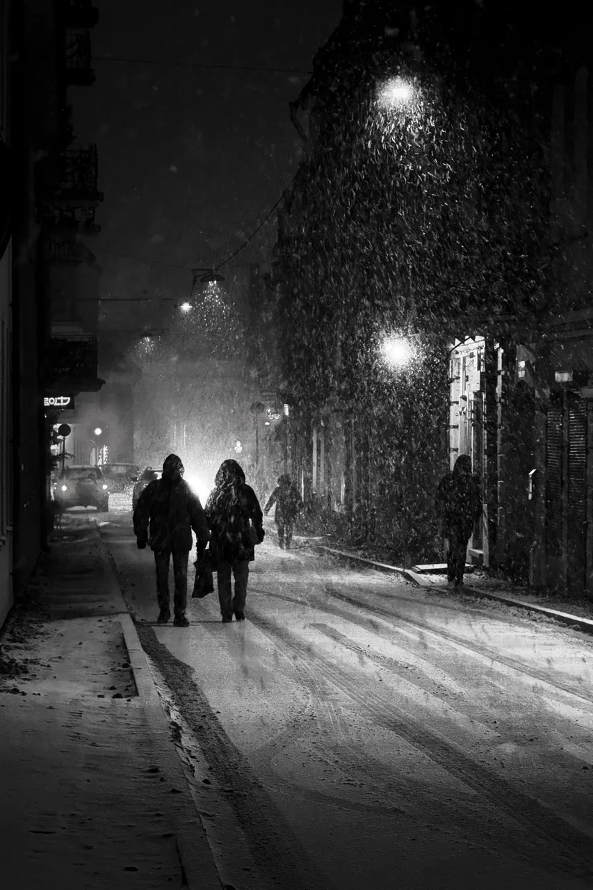 Two people, bundled in coats, walk down a snow-covered city street at night, illuminated by glowing streetlights amidst falling snow, with a partially visible store sign reading "BOLT."