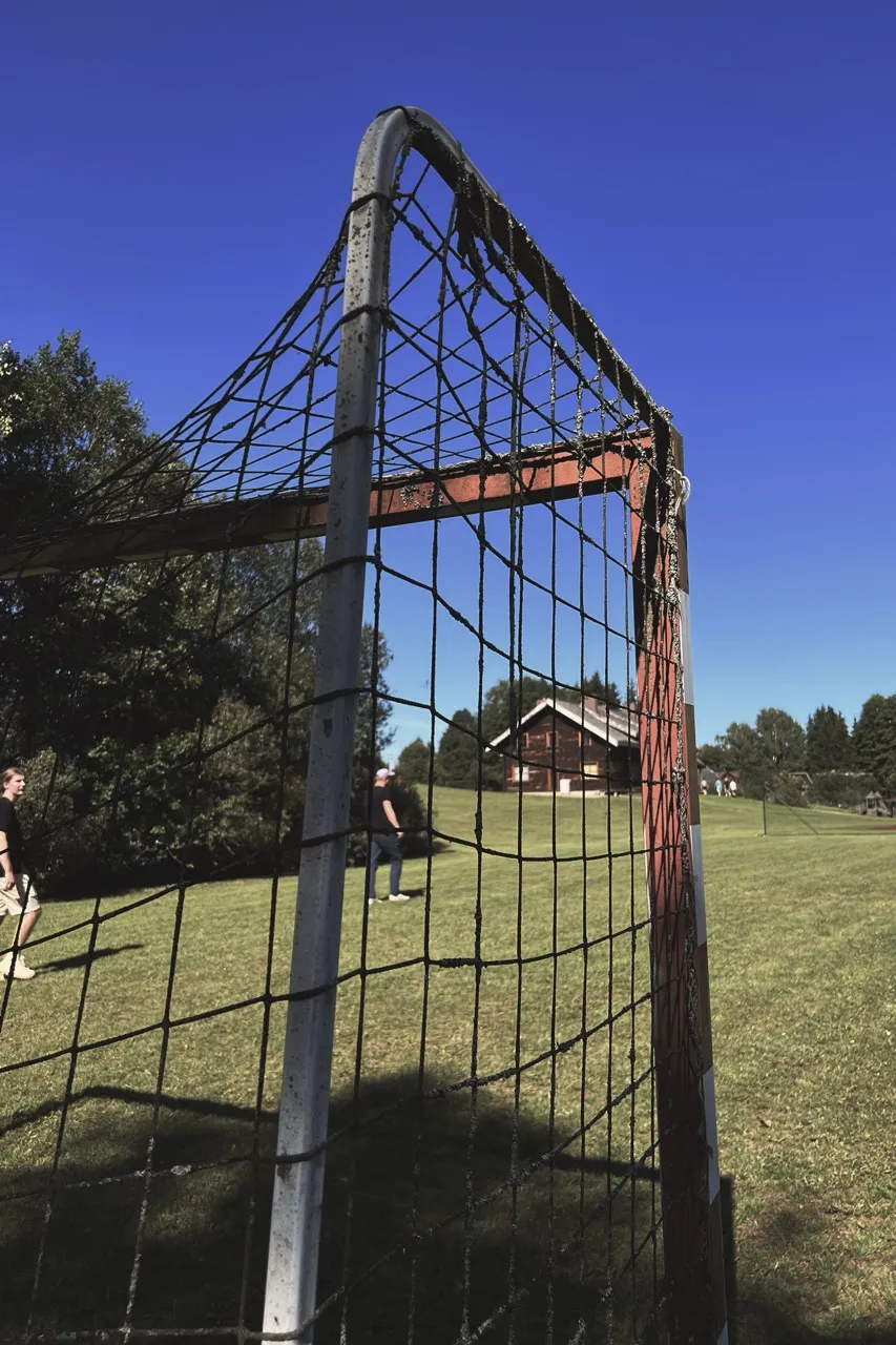 A soccer goal with a metal frame and netting stands on a grassy field. Two people walk nearby, with trees and a building in the background under a clear blue sky.